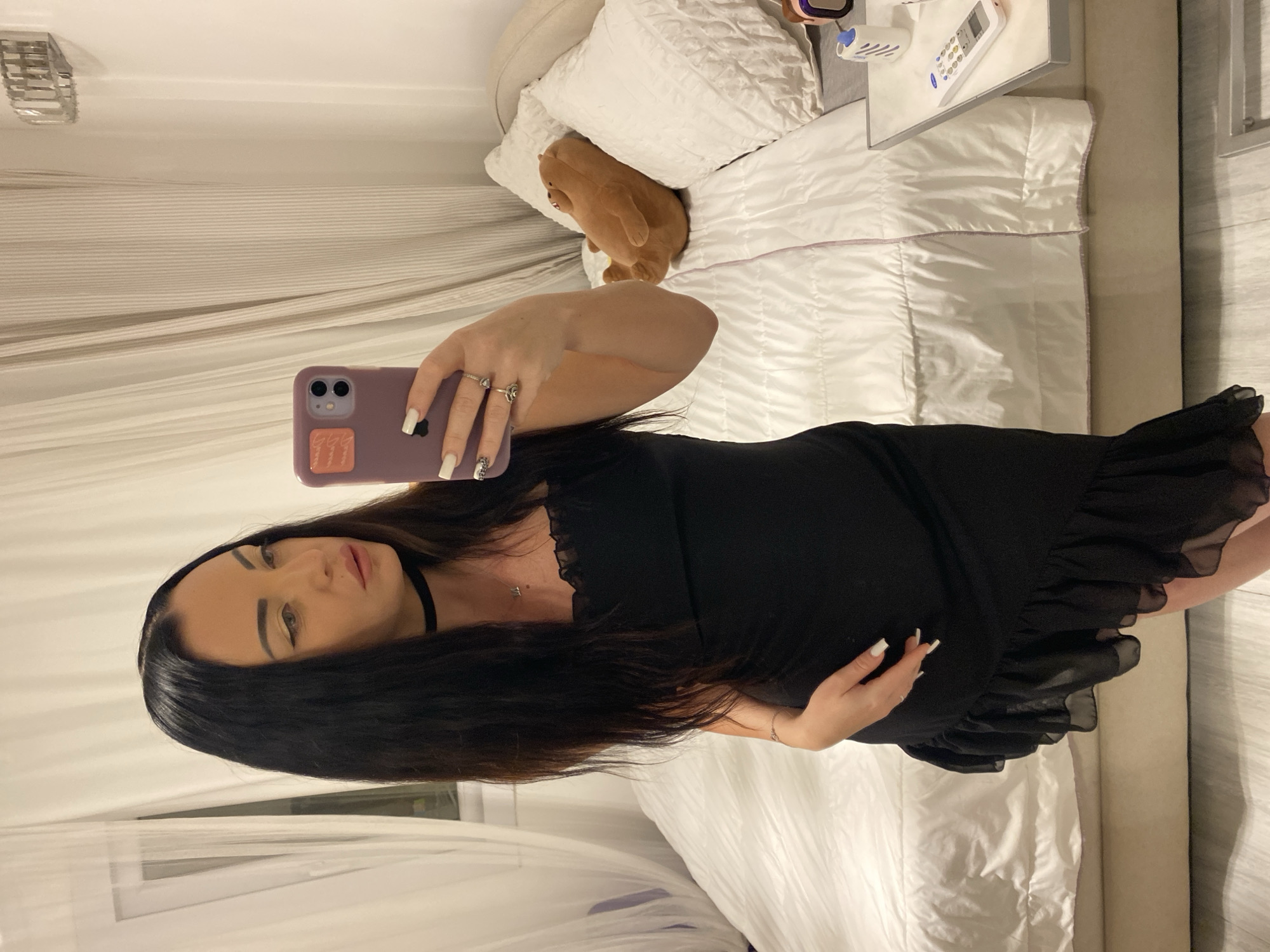 A woman stands in a bedroom, holding a smartphone to take a mirror selfie. She is wearing a black, ruffled dress and a choker necklace. Her dark hair is long and straight, and she has on winged eyeliner. The room features a large bed with white bedding and several pillows, one of which is a plush bear. A bedside table with a remote control and other items is visible. The background includes sheer white curtains and vertical striped curtains. The lighting suggests it is evening or indoors. This scene is in Sharm El Sheikh, Egypt.