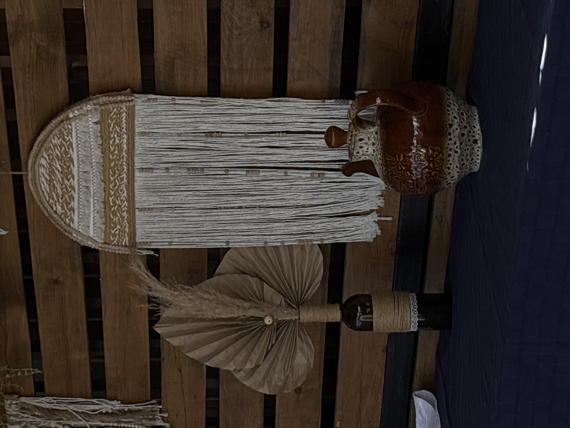 This is a rotated image of a rustic display against a wooden plank background. The display features several decorative objects. On the left, a wall hanging made of white and beige macrame and lace is visible. To its right, a brown ceramic pitcher with decorative cutouts and floral patterns sits on top of the macrame. Below the pitcher, a glass bottle wrapped in twine and lace stands upright. In front of the bottle, a paper fan decoration and a fluffy dried plant are arranged. The scene appears to be outdoors, possibly at an event or market, given the rustic aesthetic and the presence of decorative items. The lighting suggests it might be daytime. There is no visible text in the image.