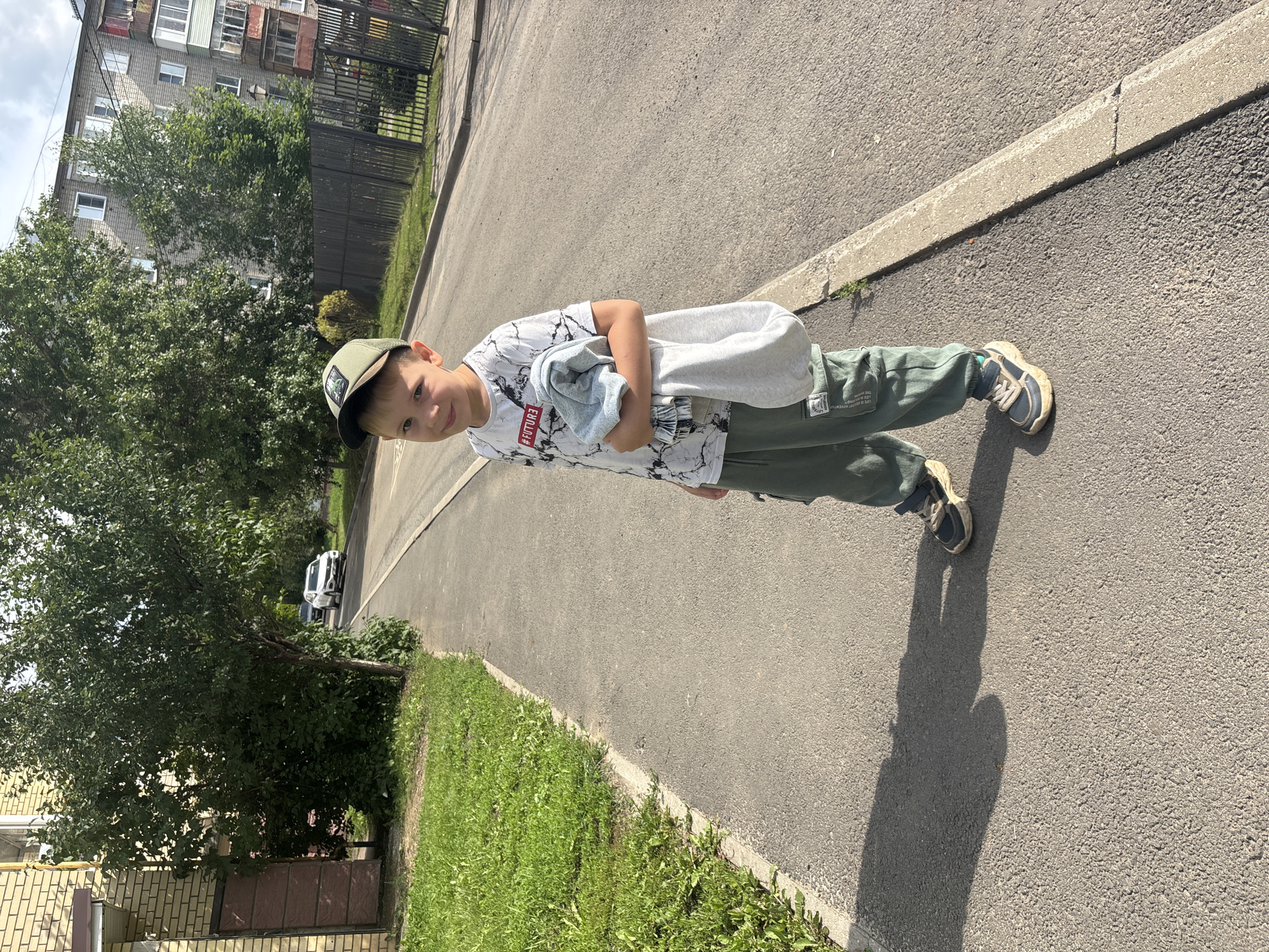 A young boy, wearing a green baseball cap, a white t-shirt with a black marble pattern and red text, and green cargo pants, stands on a paved sidewalk. He is holding a light blue and gray folded garment in his arms. The boy is looking towards the camera with a slight smile. To his left is a strip of green grass and a brick building with a brown door. To his right, the paved sidewalk transitions to a street with a white car in the distance. Behind him, there are trees and a multi-story apartment building. The scene appears to be in a residential area in Rusanovo, Russia, on a sunny day.