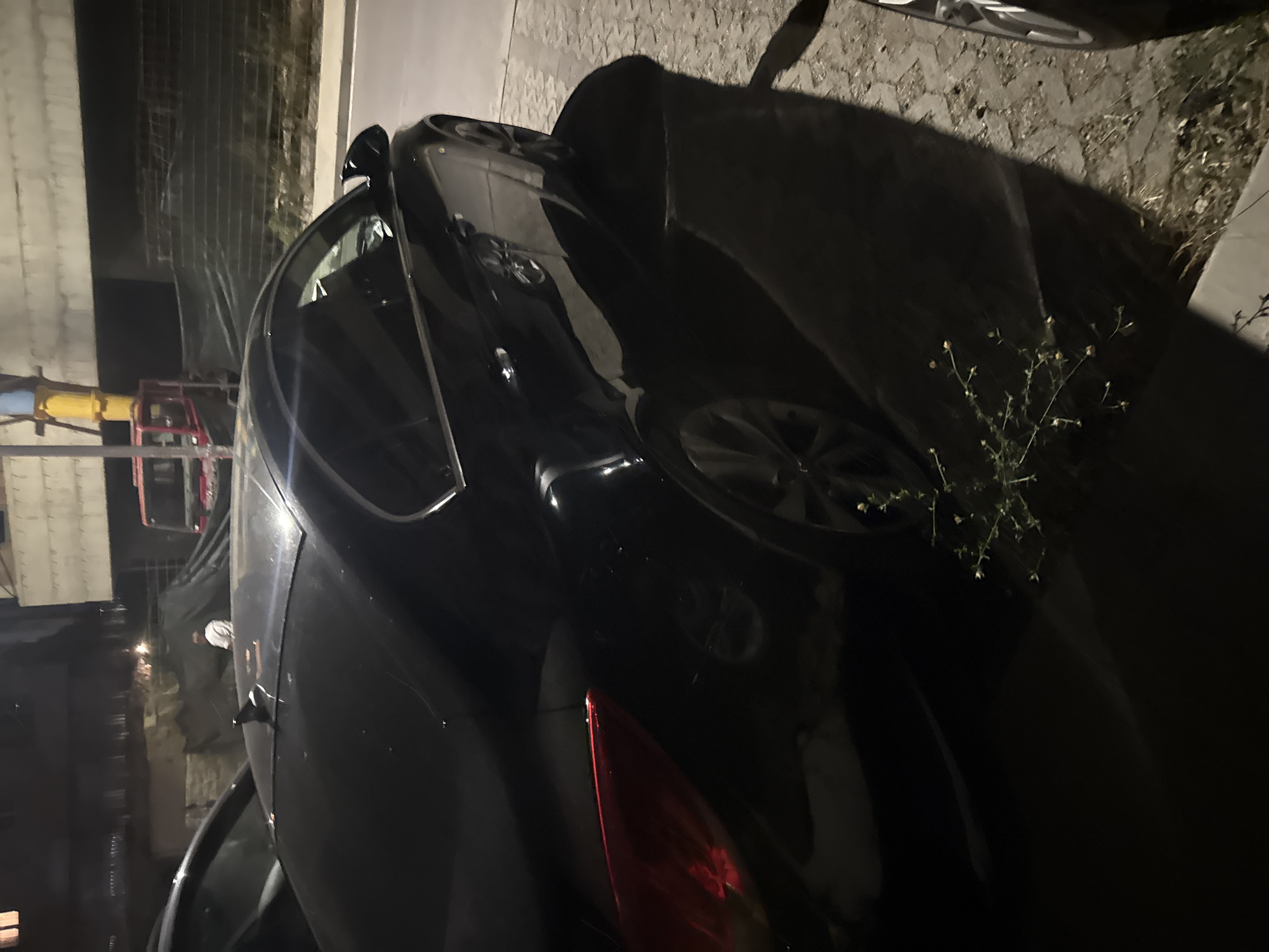 A black sedan is parked on a paved surface at night. The car is angled, showing its rear passenger side and rear. The car has a dark interior, chrome trim around the windows, and alloy wheels. A small, sparse shrub with yellow flowers is growing to the right of the car. In the background, a red truck with a crane is visible, as is a multi-story building. The area appears to be a construction site or an undeveloped area within the city of Boreti, Montenegro.