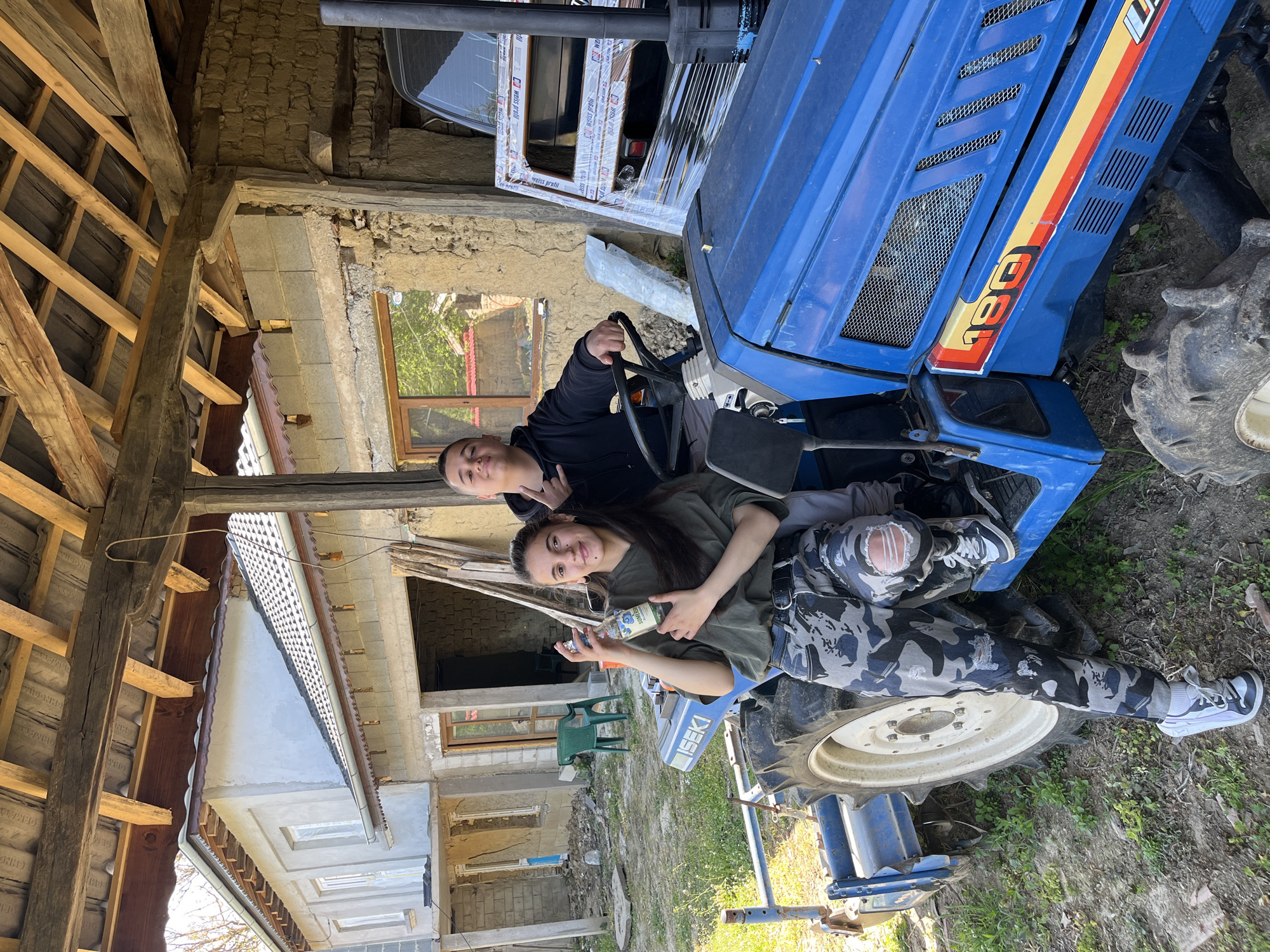 Two people are on a blue ISEKI 180 tractor. One is seated in the driver's seat while the other is reclining on the hood. They appear to be posing for a photo. The woman lying on the hood is holding an unlabeled bottle. The background includes a partially renovated building, a wooden support structure, and various construction materials. The scene is outdoors in what appears to be a rural setting. The location is Rish, Bulgaria.