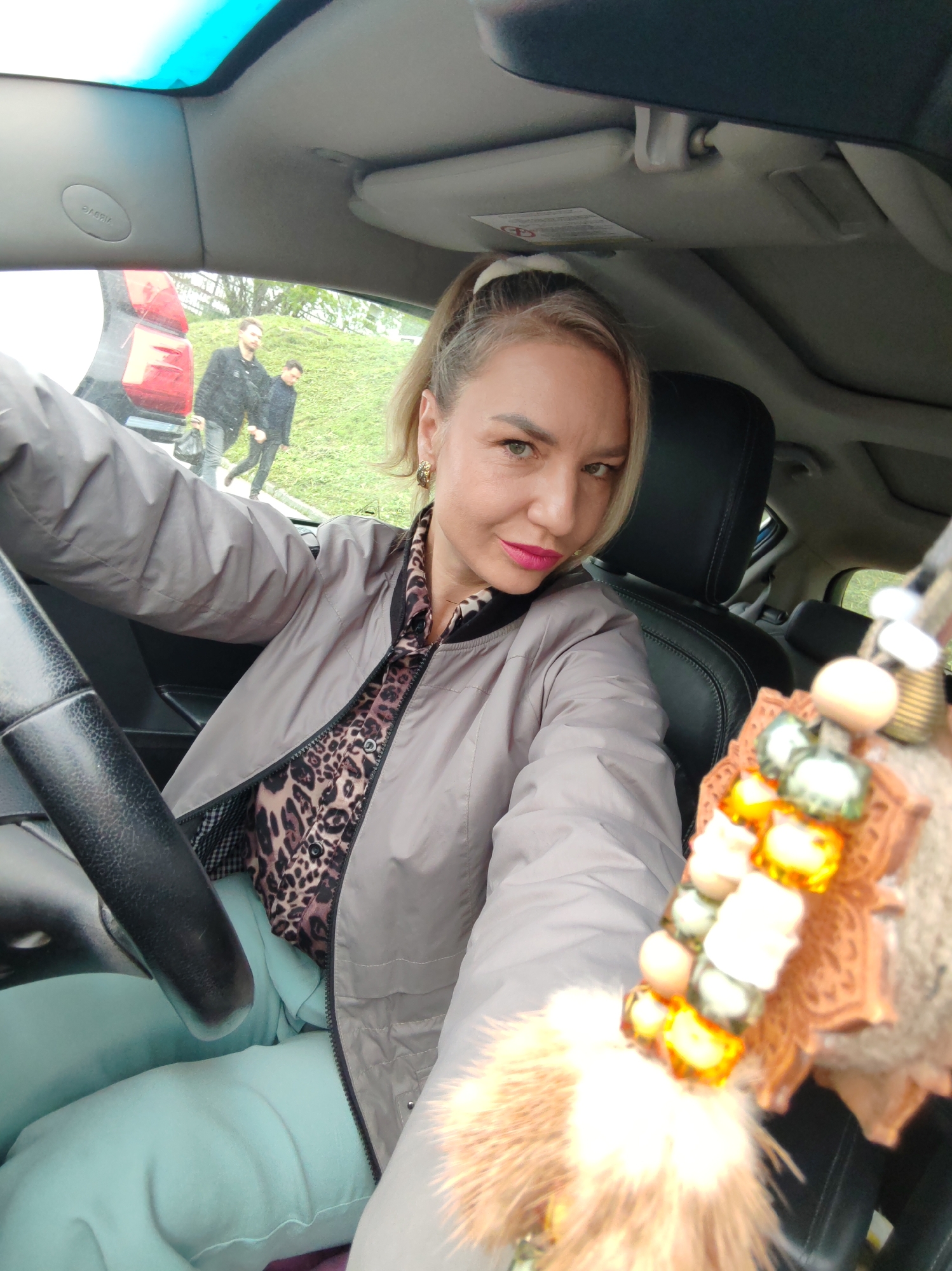 A blonde woman with bright pink lipstick and a leopard print shirt is sitting in the driver's seat of a car, taking a selfie. She is wearing a light gray bomber jacket. In the background, two men are walking away from a red car on a grassy embankment. A fuzzy keychain with beads hangs from the rearview mirror. The scene appears to be outdoors, possibly in a parking lot or on the side of a road, with some trees visible in the distance.