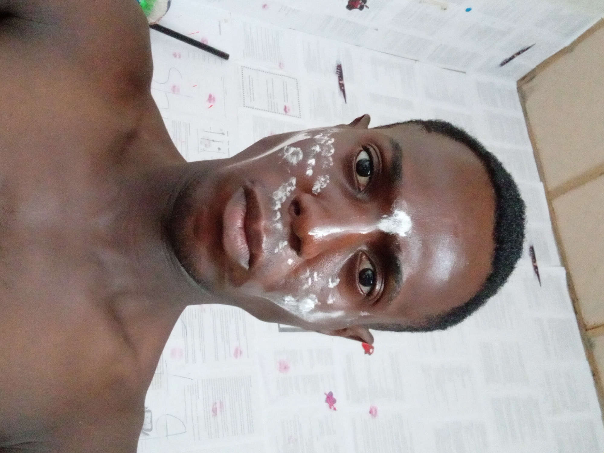 The image is a close-up selfie of a dark-skinned man with a shaved head. He has white spots of an unidentified substance on his face, primarily around his nose and forehead. His gaze is directly at the camera. The man's upper body is visible, revealing his bare shoulder. The background appears to be a wall covered in papers with text, possibly in a room. The location mentioned is Akwete, Nigeria. No specific actions or interactions are evident.