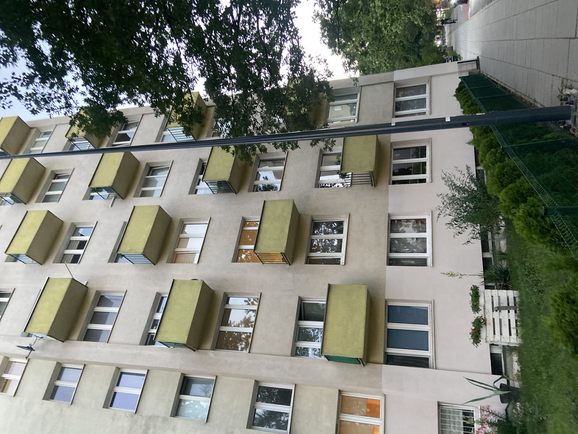 The image shows a multi-story apartment building in Warsaw, Poland, with a sidewalk and trees in the foreground. The building has a light-colored facade with numerous windows and balconies with light green railings. Several trees are visible, casting shadows on the building and sidewalk. The sidewalk is paved with concrete tiles and a green fence runs along the edge of the property. There are a few people on bicycles riding on the sidewalk in the distance. The overall scene suggests a residential area in an urban setting.