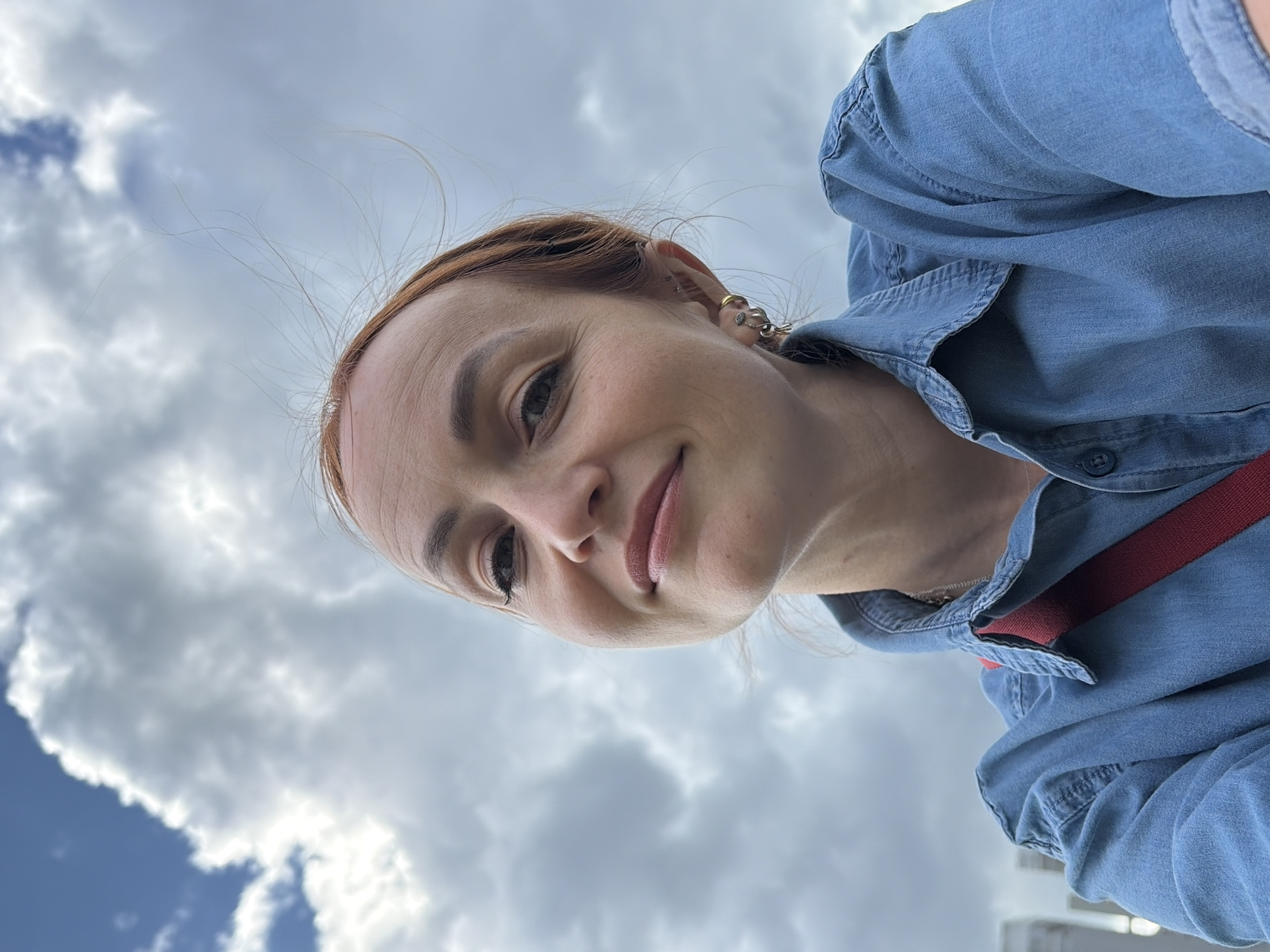 A selfie shows a Caucasian woman with auburn hair and fair skin against a cloudy sky. She is wearing a denim shirt and a red strap crosses her chest. The woman has makeup on and is smiling slightly. Her hair is windswept. The background appears to be an outdoor environment with a cloudy sky, suggesting an overcast day. There are no specific discernible landmarks or architectural features visible that would indicate the location in Chaikovsky, Russia.
