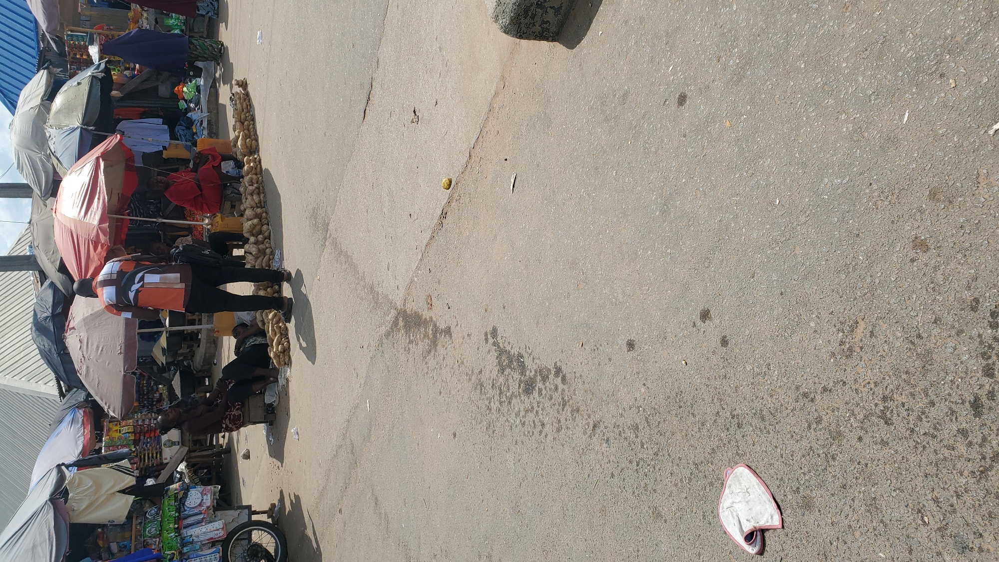 In Jalingo, Nigeria, an outdoor market scene unfolds under a bright sky. The foreground is dominated by a gray asphalt road, with scattered debris and a light-colored fabric item, possibly a cloth, lying to the left. In the midground, several market stalls are set up, covered by various umbrellas and awnings. People are present, with one individual standing and facing away from the camera, wearing an orange and white patterned shirt and black pants. Another person, possibly a vendor, sits on a wooden stool under a red umbrella, wearing a red outfit. A third person, wearing a dark headscarf and patterned skirt, is visible in the background. Piles of produce, likely yams, are neatly arranged on the ground in front of the stalls. The atmosphere suggests a typical day in a bustling Nigerian market, with goods being sold and people engaged in daily commerce.