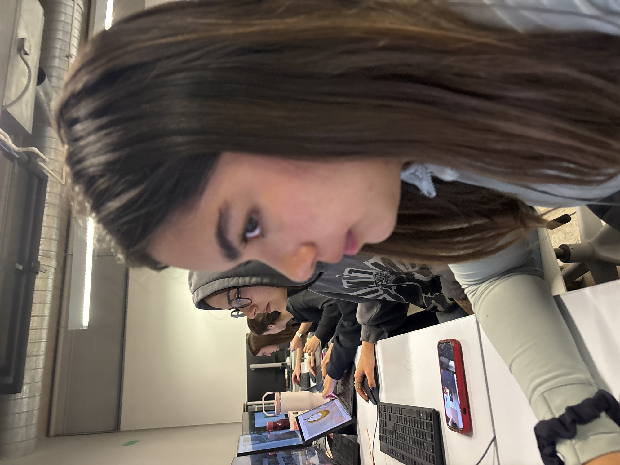 The image shows a group of people sitting at desks in a room, possibly a classroom or office. The room appears to be indoors with fluorescent lighting and white walls. The people are focused on using computers and tablets. One person in the foreground is using a mouse, while a cell phone with a red case stands on the desk beside the keyboard. The location is unknown and there is no indication in the image that it is in Mexico City, Mexico.