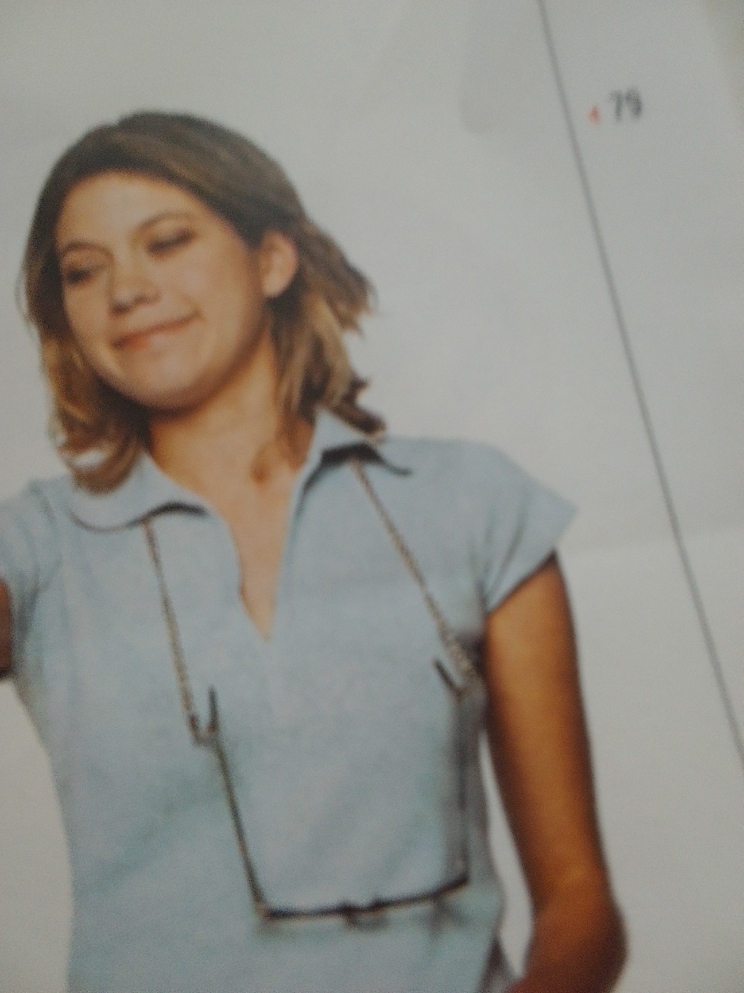 A young woman with light brown hair smiles at the camera. She is wearing a light blue collared shirt. A pair of glasses hang from a cord around her neck, resting on her shirt. Her arms are visible, with her right arm extending out of the frame. The background is a plain, light color. There is text in the upper right corner that reads "79". The image appears to be a photograph, possibly from a catalog or yearbook, taken indoors in what seems to be a studio setting. The lighting is bright and even, suggesting artificial light.