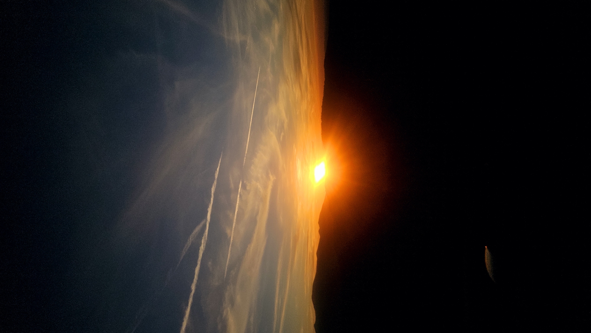 The image depicts a sunset over a mountainous landscape. The sun, a bright, intense yellow-orange orb, is setting behind a dark silhouette of hills, casting a warm glow across the sky. Wispy clouds, tinged with orange and pink, stretch across the horizon, some appearing as contrails from aircraft. The sky transitions from a deep blue at the top to vibrant oranges and yellows near the horizon. The foreground is a dark, silhouetted expanse, likely the side of a hill or mountain, with a faint, reflective object visible on the lower left. The scene is serene and evokes a sense of natural beauty and the end of the day. The location is identified as Simeonovets, Bulgaria.
