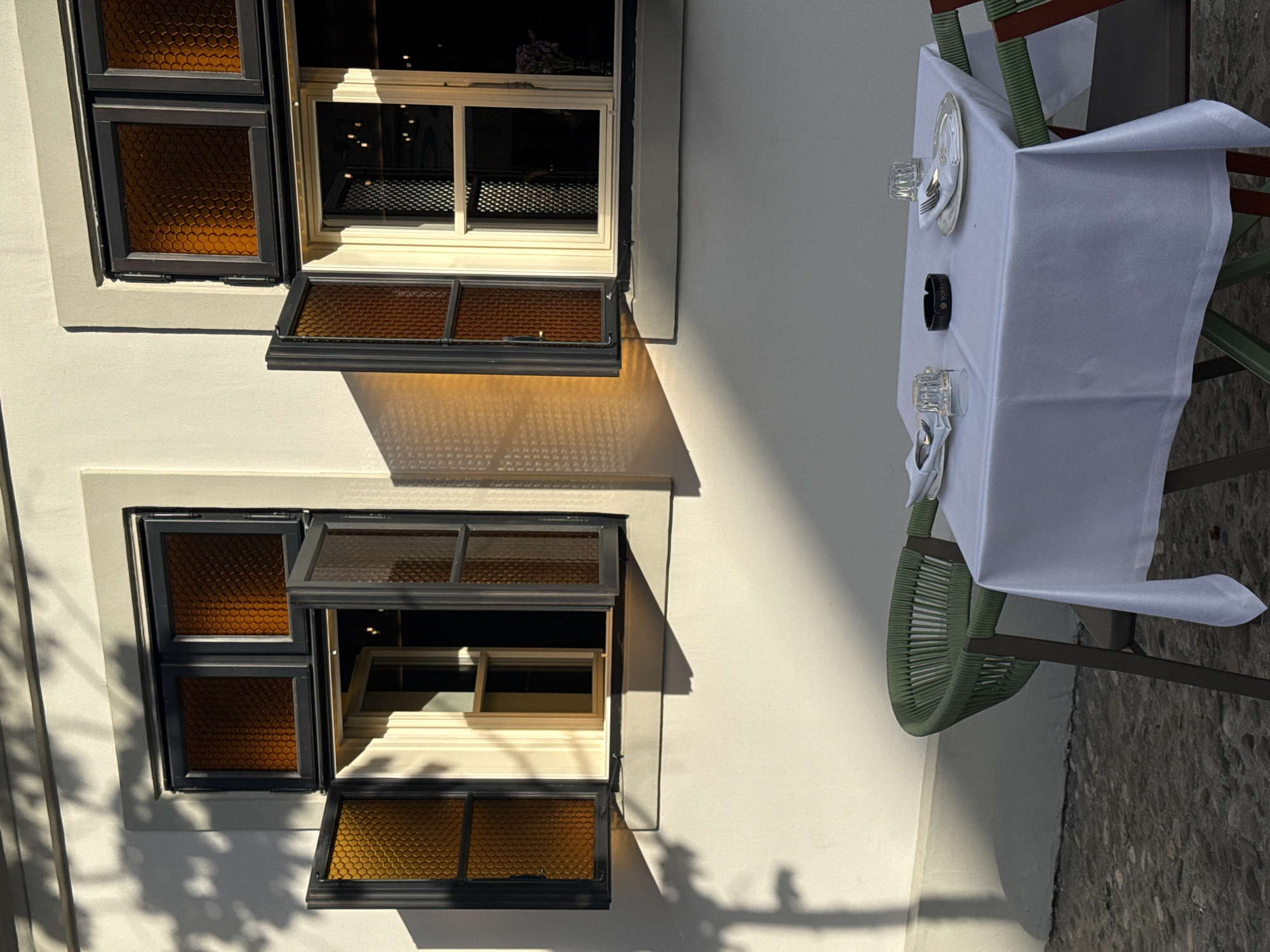 A photograph taken in Belgrade, Serbia, shows a white-walled building with two sets of honey-colored, grid-patterned windows. The lower window is partly open, and the upper window is slightly open. A table is set for two with a white tablecloth, place settings, and an ashtray. The table sits outside the building. Sunlight casts shadows on the building.