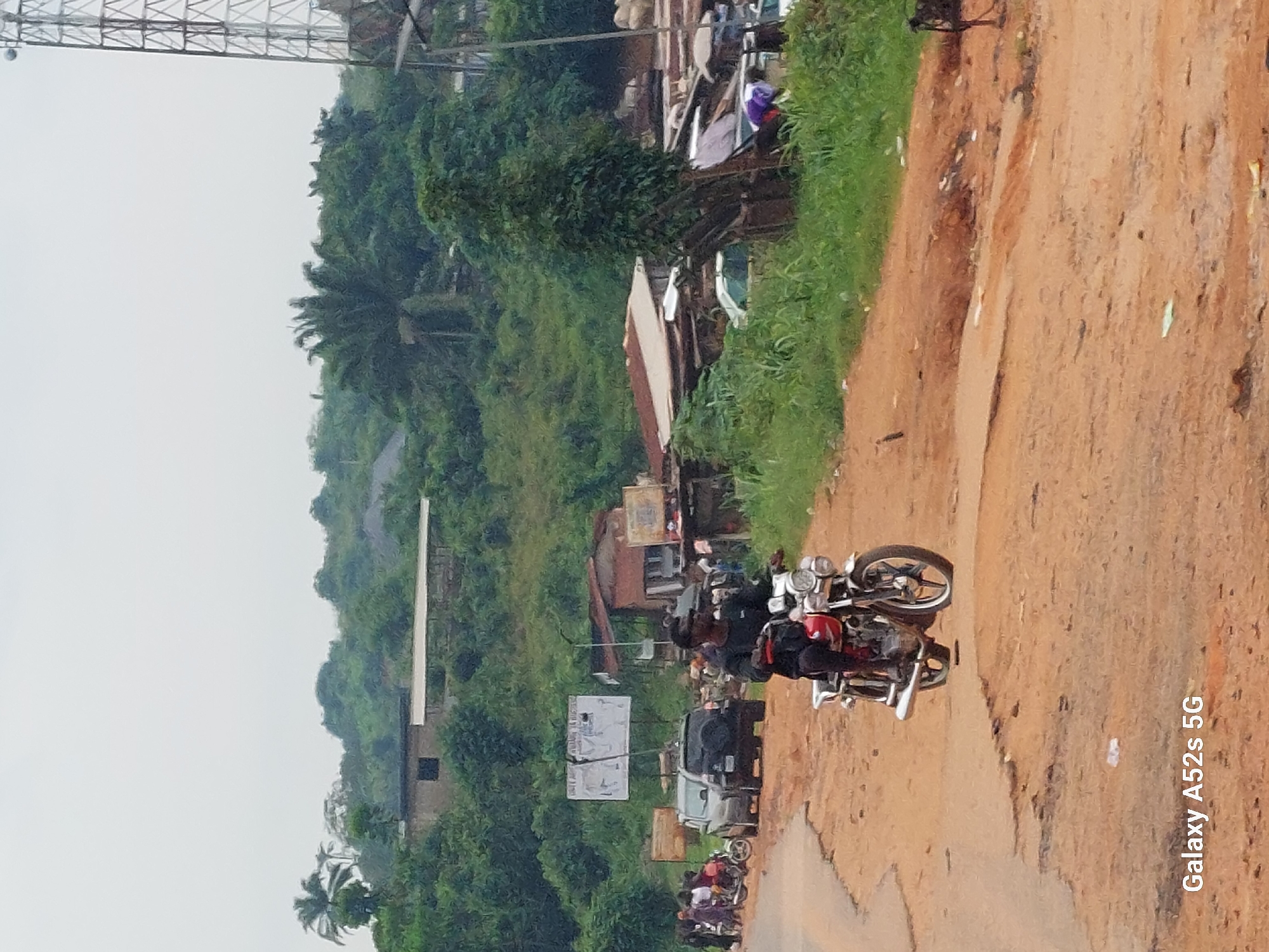 A medium shot captures a dusty, unpaved road in Awkuzu, Nigeria, under an overcast sky. A man, wearing a black long-sleeved shirt and dark pants, rides a red and silver motorcycle forward. The motorcycle has a chrome exhaust pipe and dual headlights. To the left, a few other motorcycles and several people are gathered, as are two parked vehicles: a dark SUV and a silver car. In the background, a small cluster of buildings with corrugated metal roofs is visible, situated amidst lush green vegetation and trees. Further up the hillside, more structures and dense foliage are apparent. A tall telecommunications tower stands on the right side of the frame. The road surface is rough and uneven, with patches of loose dirt and debris.