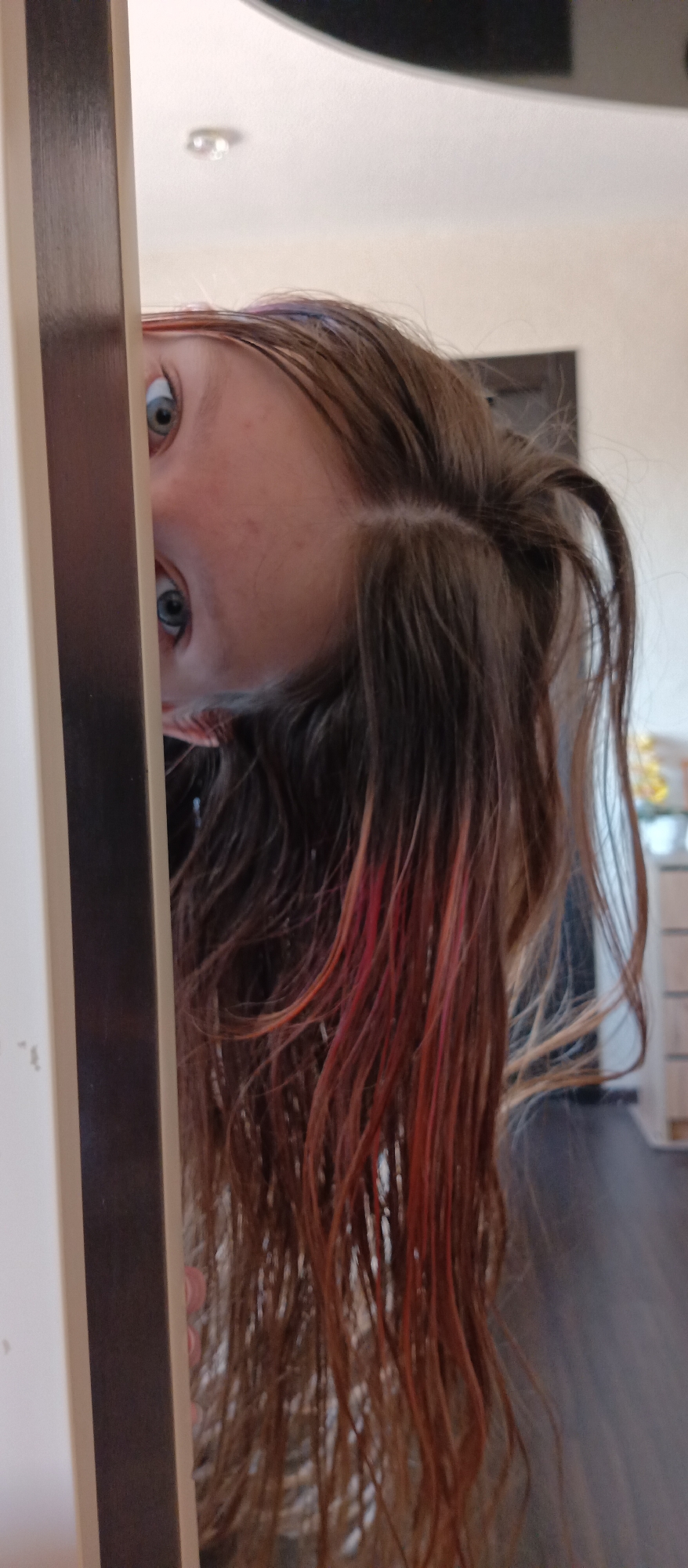 The image shows a person's face peeking from behind a door frame. The person has blue eyes and brown hair with red highlights. The background is out of focus but appears to be a room with a dresser. The location is Unknown, Unknown.