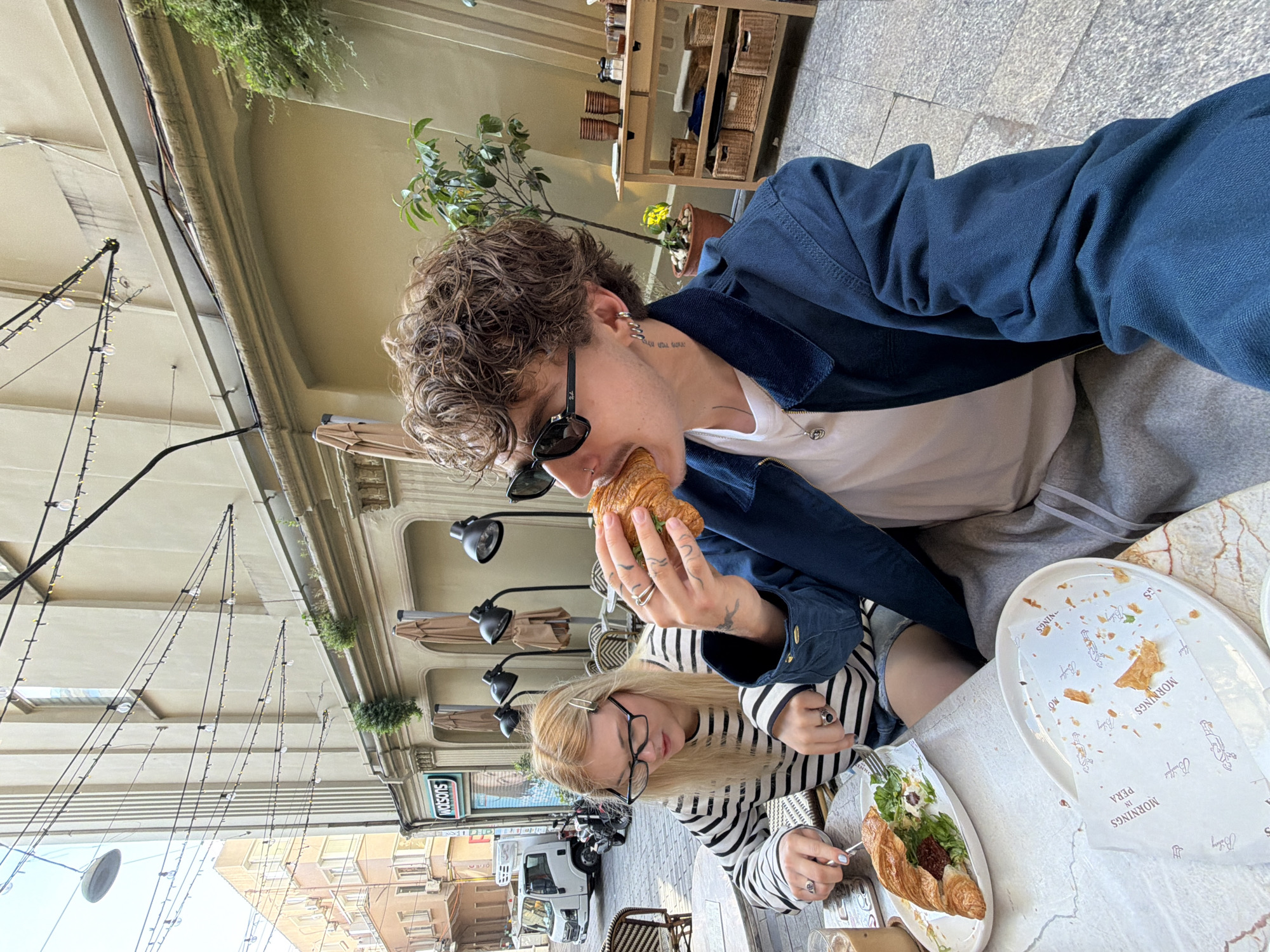 A medium shot shows a man and a woman seated at a table outdoors at a cafe in Beyoğlu, Turkey. The man, fair-skinned with curly brown hair, wears sunglasses and a blue jacket over a white shirt, and is eating a croissant. His neck is tattooed. The woman, fair-skinned with blonde hair, wears eyeglasses and a striped shirt, and is eating a croissant with a salad. They sit at a marble-topped table with plates that say "Mornings in Pera Bakery". In the background are building facades, street lamps, and an advertisement for Watsons. Overhead, lights are strung across the street.
