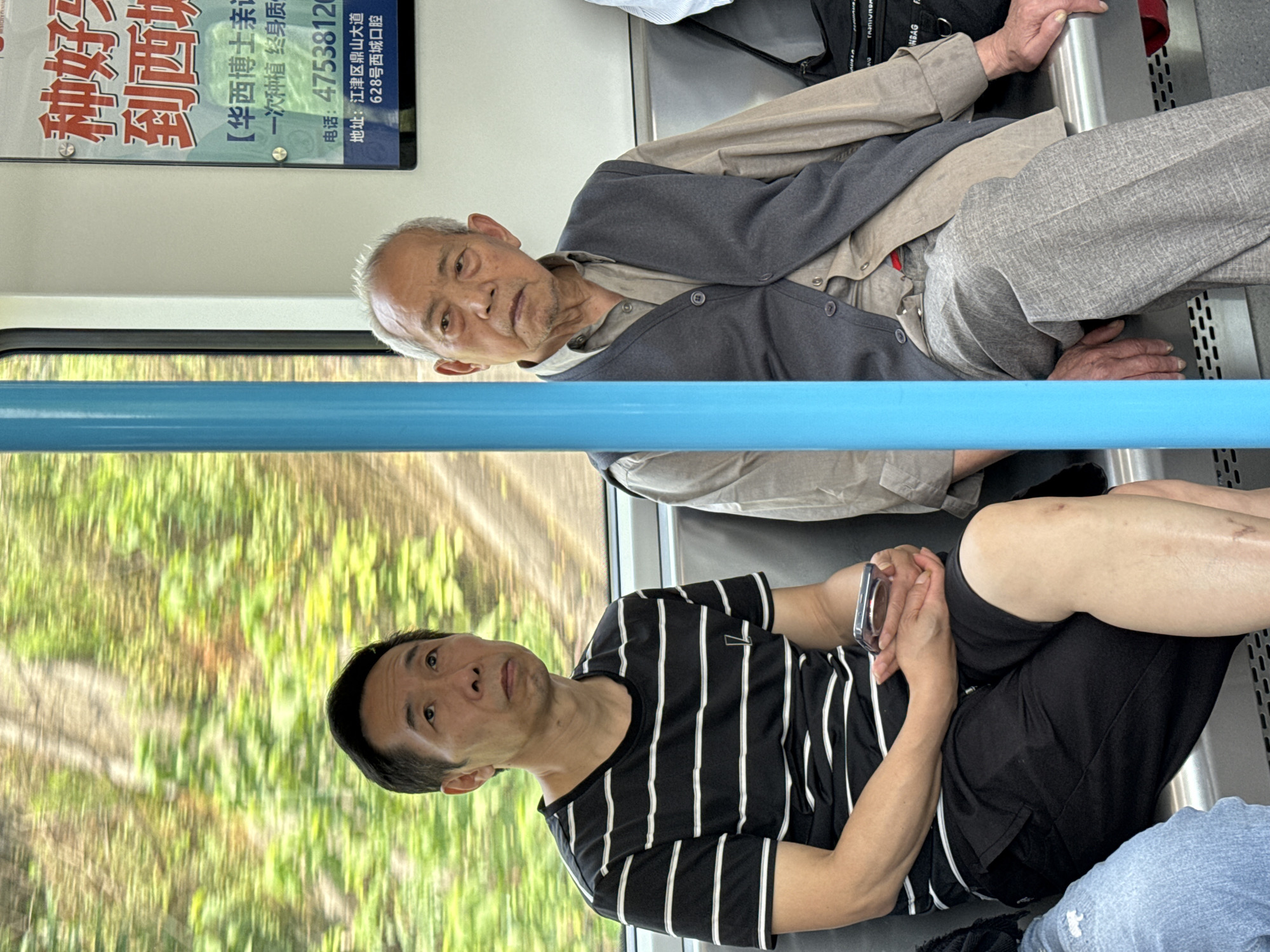 Two men are seated inside what appears to be a public transport vehicle, likely a train or subway, in Jiangjin, China. The image is taken from a slightly low angle, looking forward. A blue handrail or bar extends horizontally across the middle of the frame.
In the background, an older man with short grey hair and a goatee is seated, looking directly towards the viewer with a neutral expression. He is wearing a light-colored long-sleeved shirt under a dark grey cardigan and light grey trousers. His hands are resting on the silver frame of the seat in front of him. A dark bag is visible behind him on the seat.
In the foreground, a younger man with short black hair is seated, looking slightly to his right with a neutral expression. He wears a black t-shirt with white vertical stripes and black shorts. He holds a smartphone with both hands, his fingers intertwined, resting on his lap. Part of a third person's leg, wearing blue jeans, is visible in the bottom right corner.
Through a large window on the left side, a blurred green landscape suggests movement, with foliage and trees passing by. In the upper left corner, an advertisement poster is displayed. The text on the poster, in Chinese, reads "种好 到西城" (Plant well / Good planting to Xicheng), followed by "【华西博士亲记 一次种植终身质" (Huaxi Doctor's personal record, one-time planting, lifelong quality). Below this, contact information is provided: "电话:47538120" (Phone: 47538120) and "地址:江津区鼎山大道 628号西城口腔" (Address: Dingshan Avenue, Jiangjin District, No. 628 Xicheng Stomatology / Dental). The overall scene depicts commuters during daytime.