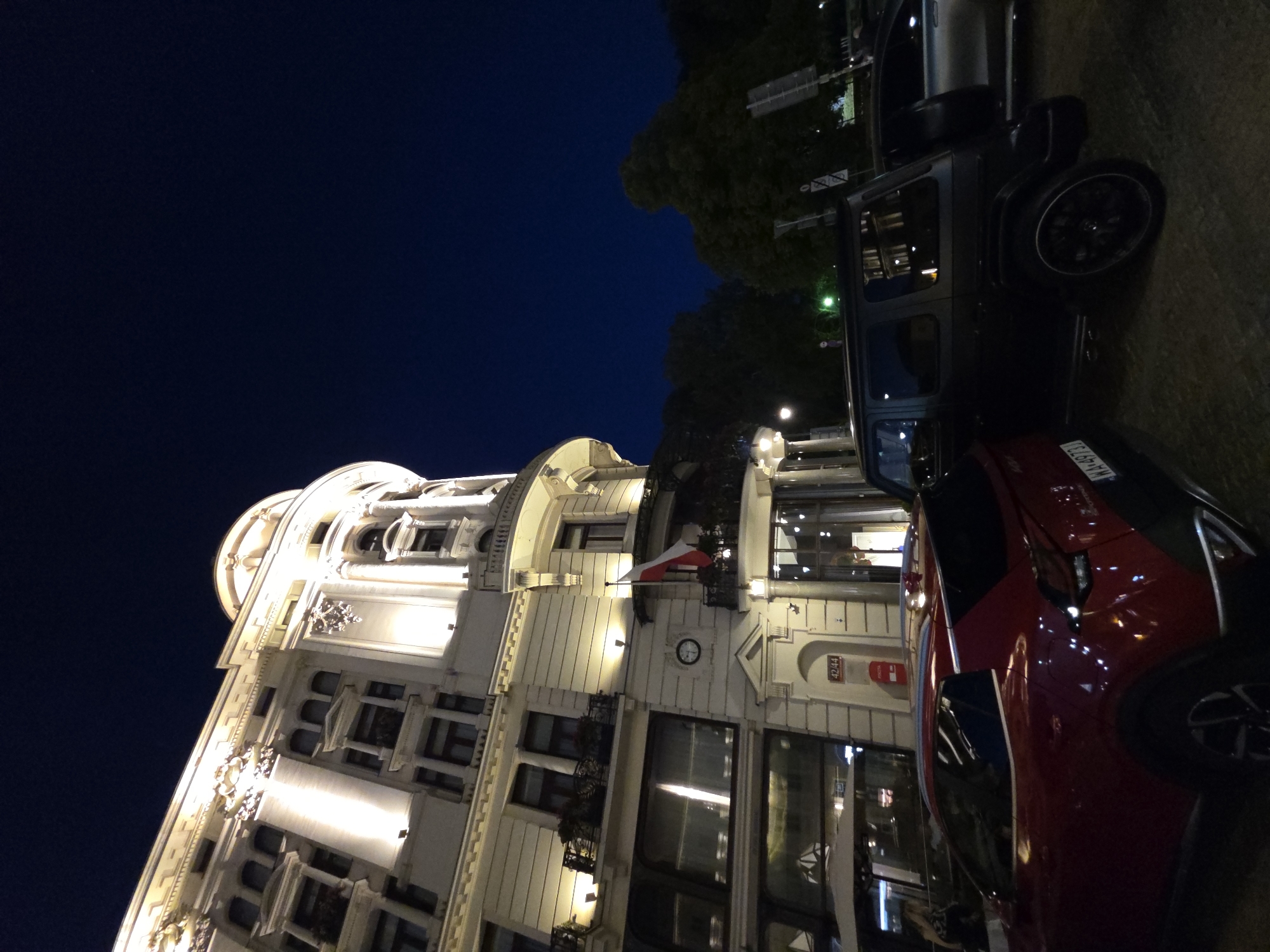 The image captures a nighttime scene in Warsaw, Poland. Prominently featured is a multi-story building with ornate, white architecture, illuminated by strategically placed lights. A Polish flag hangs from a balcony. A red car and a black SUV are parked adjacent to the building. The sky is dark blue. Street lights are visible in the distance.
