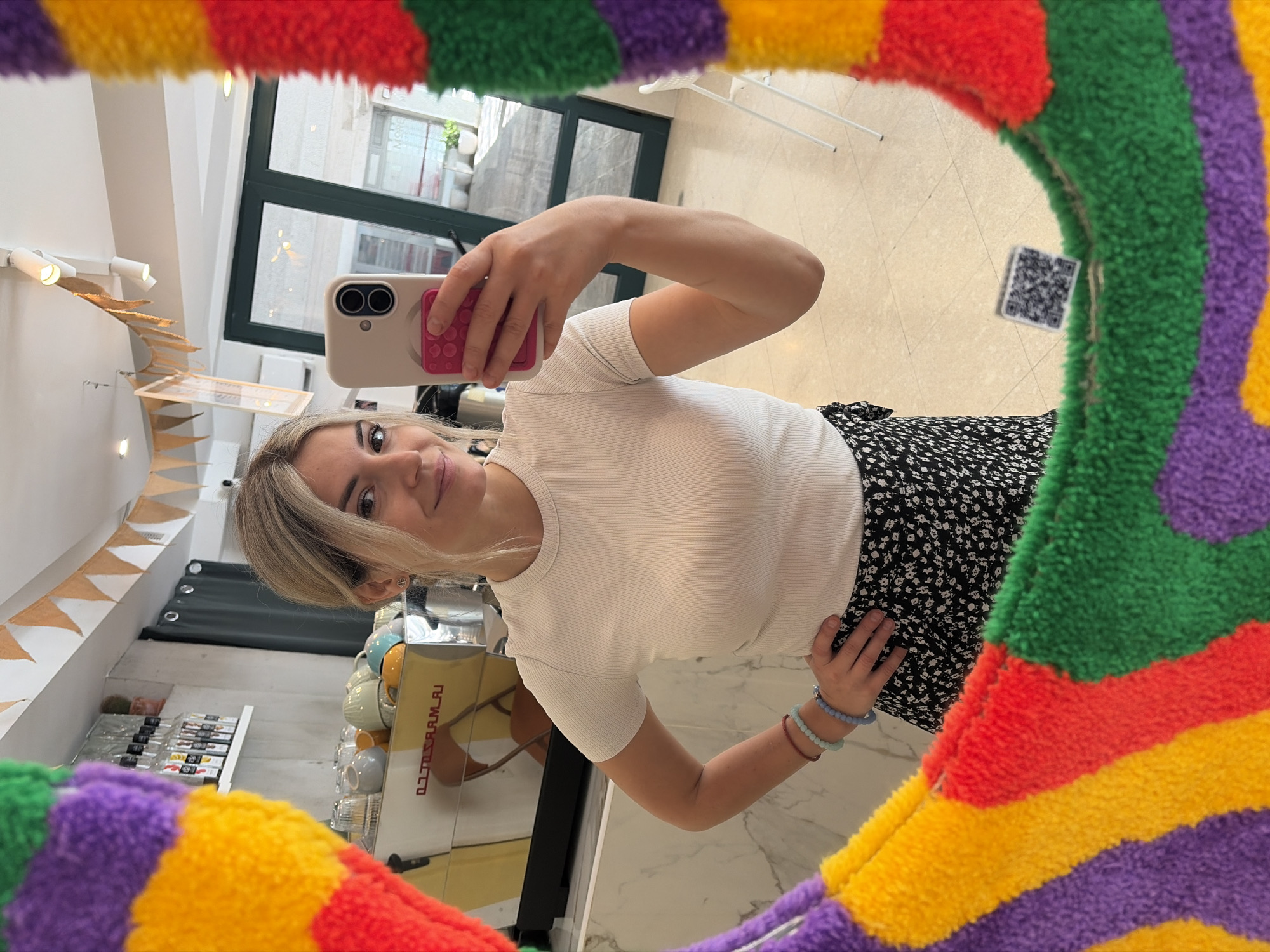 A young woman with blonde hair tied back is taking a selfie in a cafe. She is wearing a white ribbed t-shirt and a black and white floral skirt. She has several bracelets on her left wrist. In the background, there is a coffee machine and shelves stocked with bottles. A colorful, fluffy mirror frame surrounds the image. The scene is set in a cafe located in Budva, Montenegro, as indicated by the presence of a QR code on the mirror frame, which likely links to the cafe's information or location.