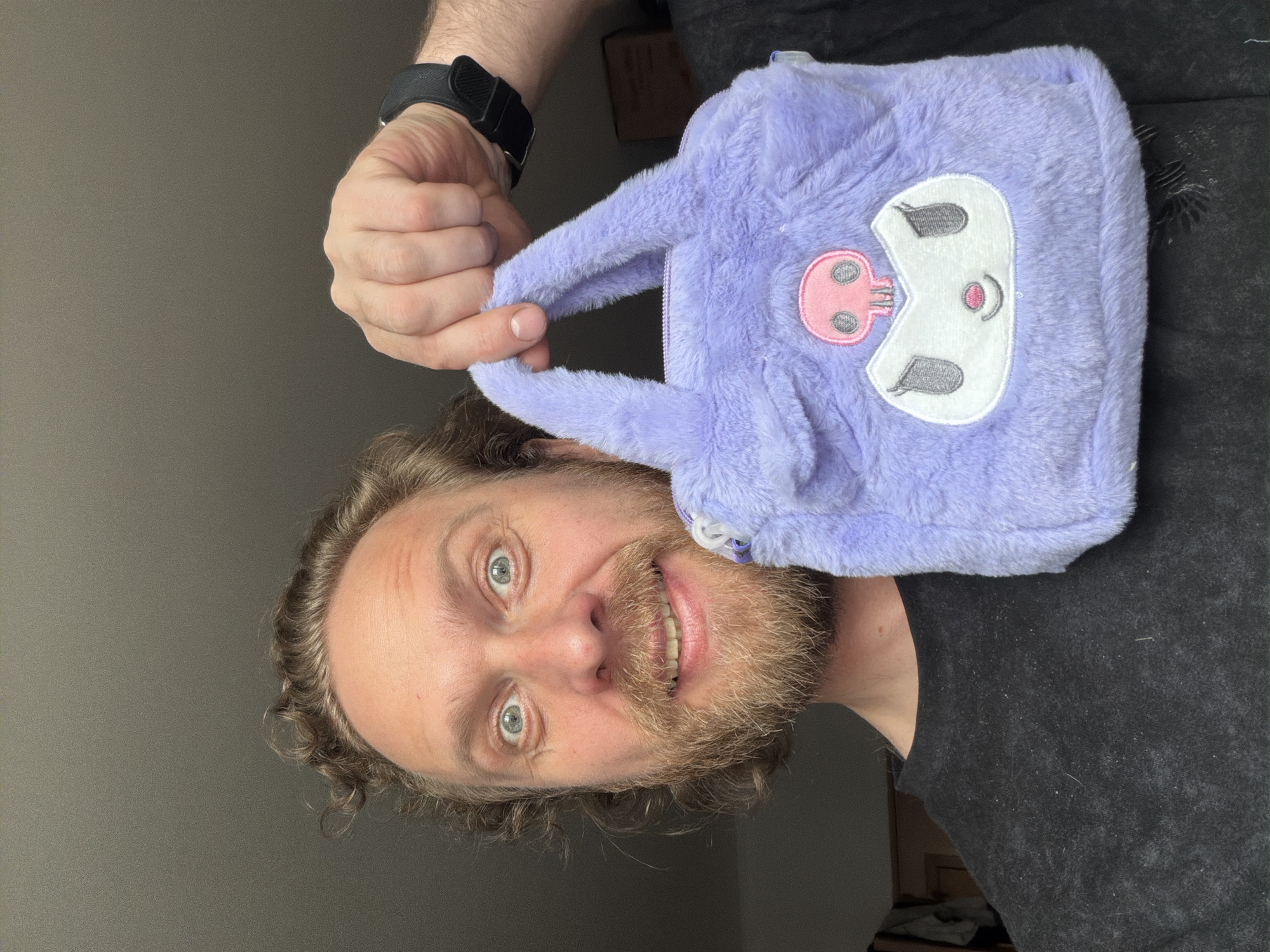 A man with light hair and a beard, wearing a black t-shirt, holds a light purple, fuzzy purse featuring a cartoonish, white and pink skeleton face. The man is smiling at the camera. He is also wearing a black smart watch on his left wrist. The background is plain and appears to be an interior setting. The location in the city of Saint Petersburg, Russia is not evident in the image.