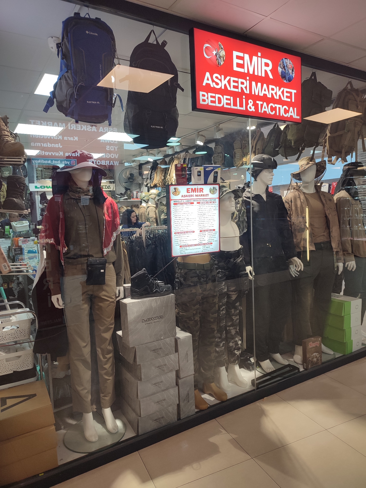 The image shows the storefront window of "EMIR ASKERİ MARKET BEDELLİ & TACTICAL" in Kadıköy, Turkey. The store displays military and tactical gear. In the foreground, mannequins are dressed in camouflage clothing and military-style hats. One mannequin wears a red scarf. To the right, other mannequins are visible in similar attire. Various backpacks, boxes labeled "LOWA," and other products are displayed inside the store window and on shelves. A red illuminated sign with white text and images of soldiers hangs above the display. The reflection of objects and people outside the store is visible on the glass. The floor outside the store appears to be tiled.