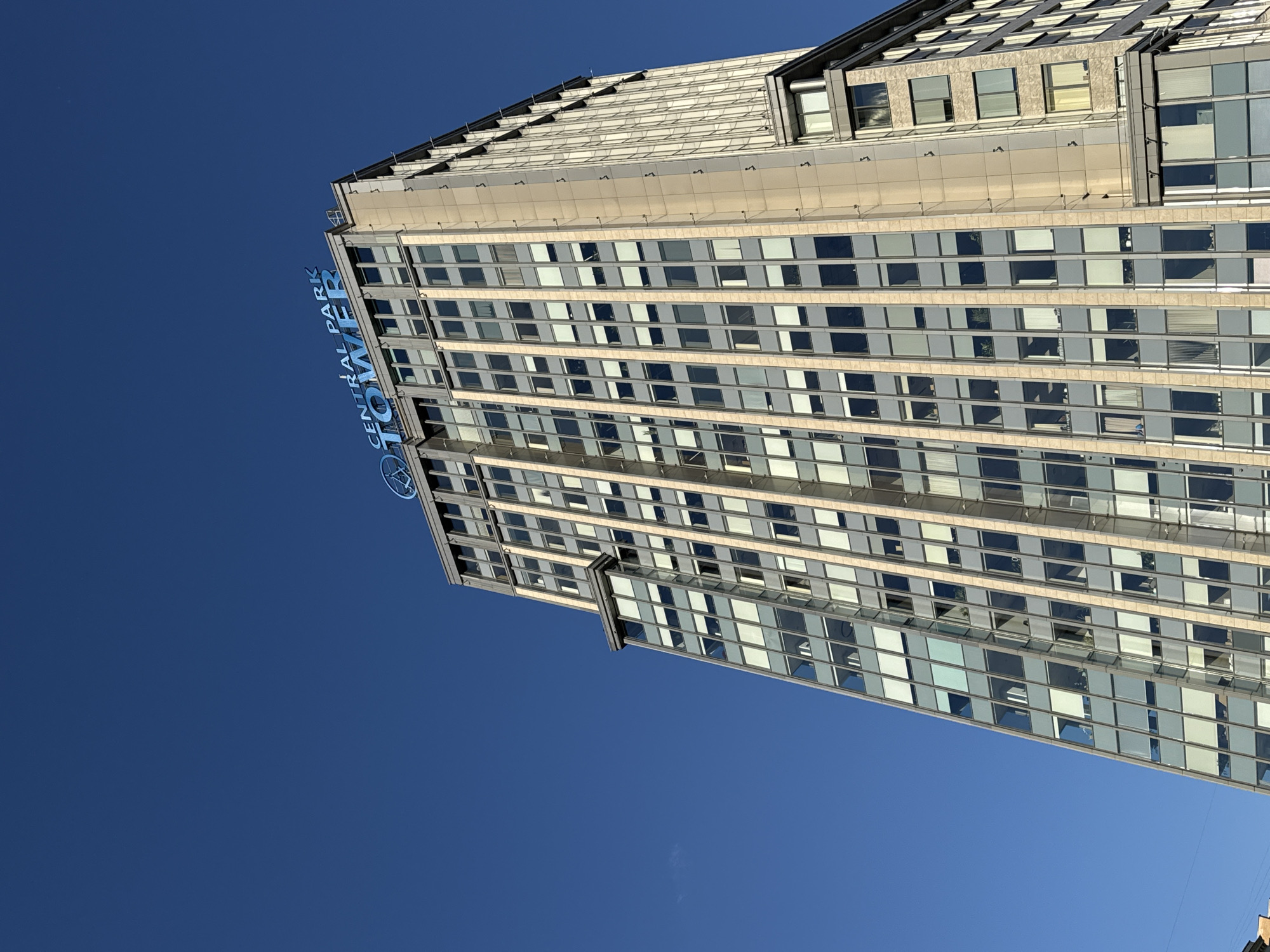 This low-angle, upward-looking shot captures the upper portion of a modern skyscraper, identified by its blue illuminated sign as "CENTRAL PARK TOWER," against a clear, deep blue sky. The building is located in Moscow, Russia, specifically the Central Park Tower business center and residential complex.
The facade is characterized by a grid pattern of numerous rectangular windows, many displaying internal blinds or curtains, set within a framework of light-colored vertical and horizontal panels, possibly stone or metal. The main visible side of the building features alternating vertical sections of light-colored material and darker grey-blue window sections. To the left of the "CENTRAL PARK TOWER" sign, a circular blue logo with an abstract design is affixed to the roofline. The building exhibits multiple architectural elements, including a prominent horizontal protrusion on its left side and a slightly different facade treatment on its right-hand corner, which appears to be clad in lighter, possibly textured panels. A tiny, faint wispy cloud is discernible in the far upper-left corner of the sky. No individuals or active subjects are visible in the frame; the scene is static and devoid of human presence. The bright, even lighting suggests a clear, sunny day.