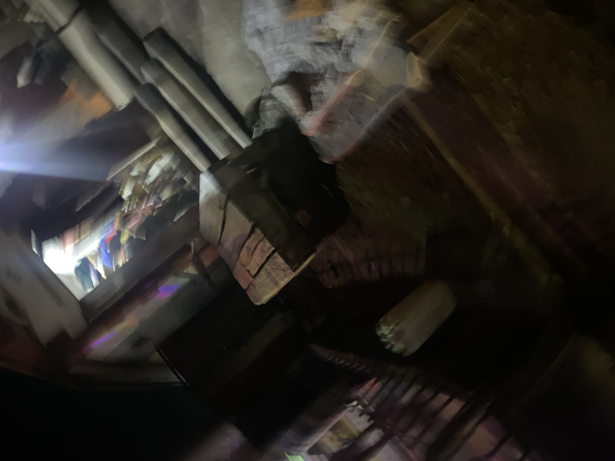 The image depicts a dimly lit, possibly indoor, scene with a strong focus on structural elements and what appears to be stored items. The perspective is angled, looking upwards or into a complex arrangement of beams and shelves. Several thick, rectangular beams run diagonally across the upper portion of the image, some appearing to be made of wood. Below these beams, a variety of objects are visible, arranged on shelves or stacked. These objects are blurred but suggest a collection of colorful items, possibly clothing or textiles, with a bright light source emanating from the left, casting a strong glare.
A prominent object in the mid-ground is a rectangular structure with a brick-like texture, possibly a beam or a decorative element, positioned at an angle. To the right of this, a cylindrical object with a textured surface and some green discoloration is visible, partially obscured by darkness. The overall impression is of a storage area, perhaps a workshop or a cluttered marketplace stall, with a sense of depth and an abundance of items.
The lighting is uneven, with bright highlights and deep shadows, suggesting artificial light and possibly a low ambient light condition. The blurriness of the image indicates movement during the capture, or a shallow depth of field. No people are clearly discernible in the frame. The setting appears to be man-made, with visible construction materials. No text is legible in the image. The context of Jos, Nigeria, suggests a warm climate, though this cannot be inferred from the visual information alone.