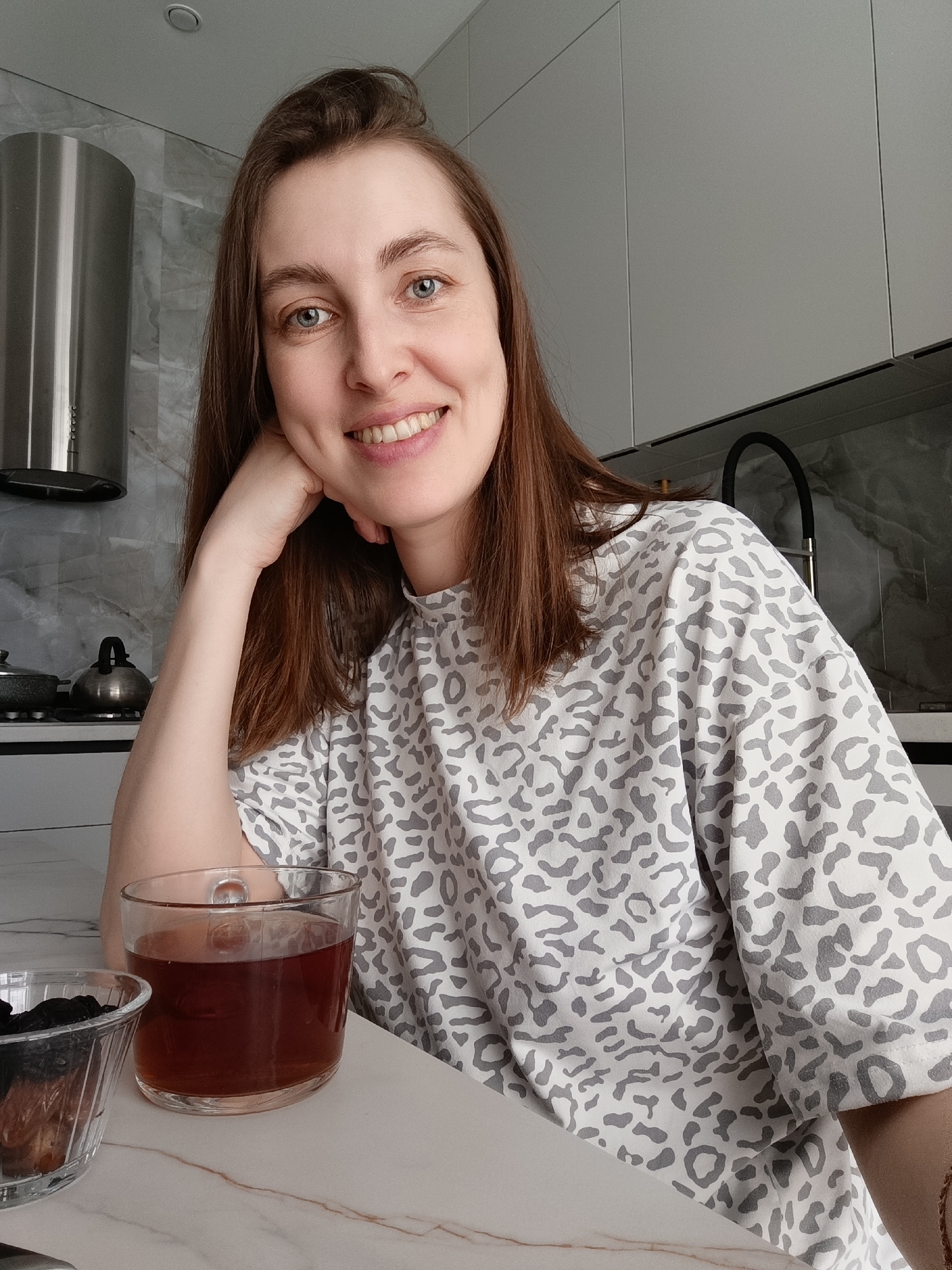 An adult female is depicted in a close-up, chest-up shot within an indoor kitchen environment. Her precise location within the city of Chaikovsky, Russia, cannot be determined from the image.
**Subject:**
A female with fair skin, brown hair, and blue eyes. She is smiling, revealing teeth, and looking directly towards the camera. Her left hand is positioned with her fingers resting against her jawline. She is wearing a light-colored (likely white or very light grey) t-shirt with an all-over grey leopard print pattern.
**Objects and Environment:**
The woman is positioned in front of a light-colored countertop with subtle brown/beige veining. On the countertop in the foreground, to her left, is a clear, ribbed glass bowl containing dark, dried fruit (consistent with prunes). To her right, on the countertop, is a clear glass mug with a handle, filled with a dark reddish-brown liquid.
The background reveals a modern kitchen setup. On the left side, a cylindrical stainless steel range hood is visible above a stovetop, where a silver metallic kettle and a dark pot are present. The wall behind the stovetop features light grey tiles with a subtle veining pattern. On the right side, a black gooseneck faucet is visible, indicative of a sink area. Above the sink area and to the right, light grey, flat-panel kitchen cabinets are installed. The ceiling is light-colored and includes a recessed light fixture.