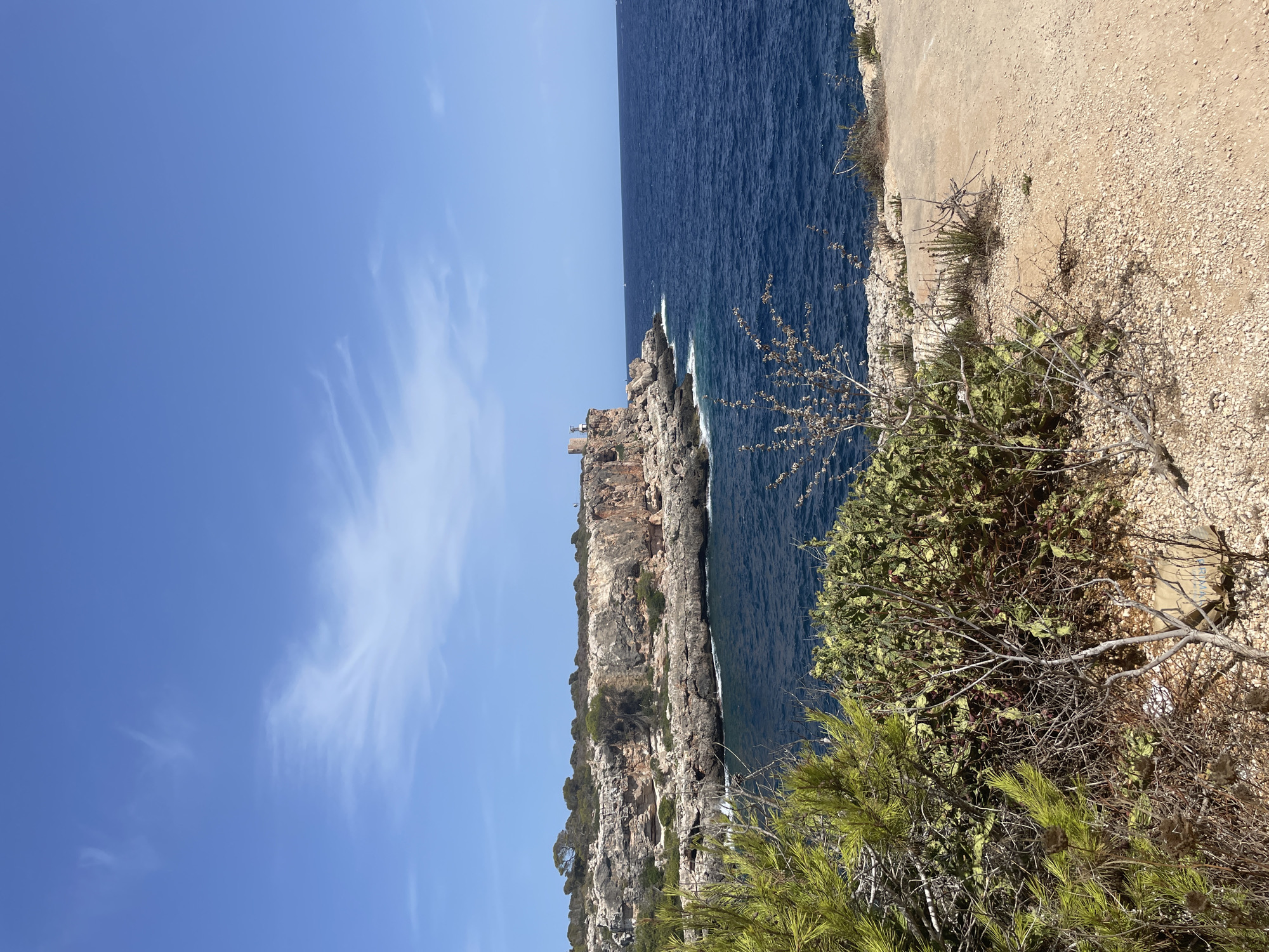 The image shows a coastal view on a sunny day. In the foreground, there is dry brush and gravel. Behind the brush, a rugged cliff face lines the deep blue sea. The cliff is dotted with sparse vegetation and rocks. Further along the coast, a small, square stone tower or structure stands on the edge of the cliff. The sky is a clear blue with a few wispy white clouds. The location appears to be a natural, unpopulated area. This scenery is consistent with coastal areas in the municipality of Santanyí, Spain, particularly the eastern coastline which features dramatic cliffs and coves.