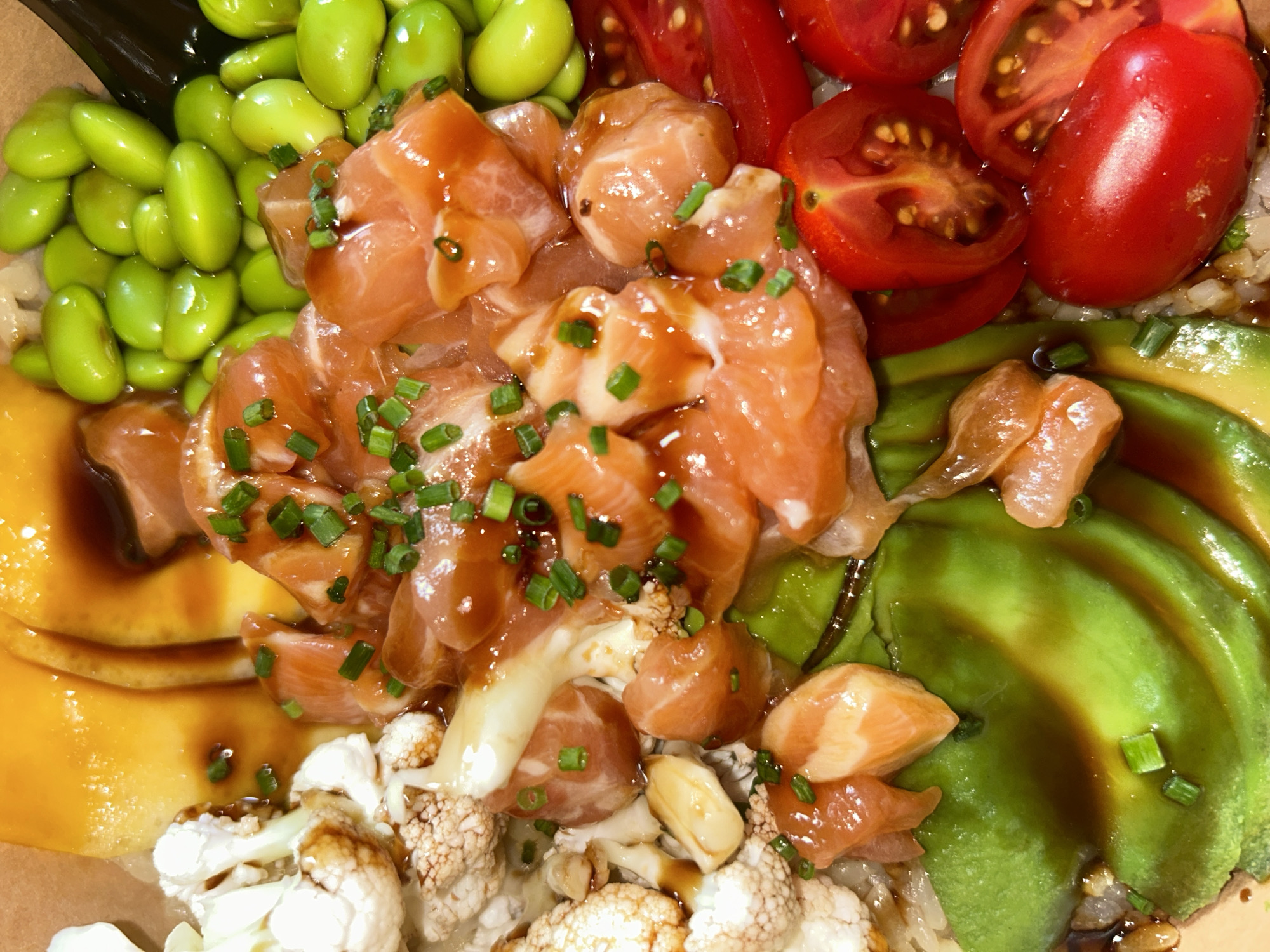 This is a close-up, overhead shot of a colorful poke bowl. The bowl is filled with various ingredients, including cubes of raw salmon marinated in a glossy sauce and sprinkled with chopped chives. Other components visible are bright green edamame beans, sliced cherry tomatoes, creamy avocado slices, and florets of cauliflower. The dish is presented in a brown paper bowl, suggesting a casual dining or takeout setting.
The lighting is bright and appears to be natural, creating a slight sheen on the food. The colors are vibrant and appealing, highlighting the freshness of the ingredients. There is no visible text in the image. The close-up perspective focuses entirely on the food, emphasizing its texture and arrangement. The scene is likely indoors, given the lighting and the presentation of the food. The dish appears to be prepared and ready to be eaten. Given the typical ingredients, this is likely a meal.