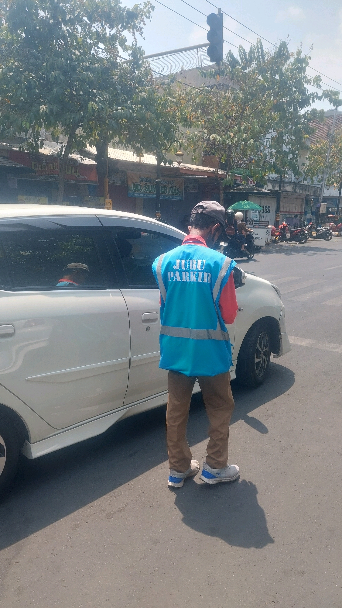 A male parking attendant wearing a light blue vest with reflective stripes over a red shirt and brown pants stands next to a white car. The vest has "JURU PARKIR" printed in blue on the back. He is wearing a dark baseball cap and a face mask. The car is parked on an asphalt road, likely in a commercial area, given the presence of shops and buildings in the background. A traffic light is visible overhead. There are also other vehicles, including motorcycles, in the background, indicating a busy street. The scene suggests an urban environment, possibly in Madiun, Indonesia, given the context provided. The overall lighting indicates it is daytime, with sunlight casting shadows on the ground.