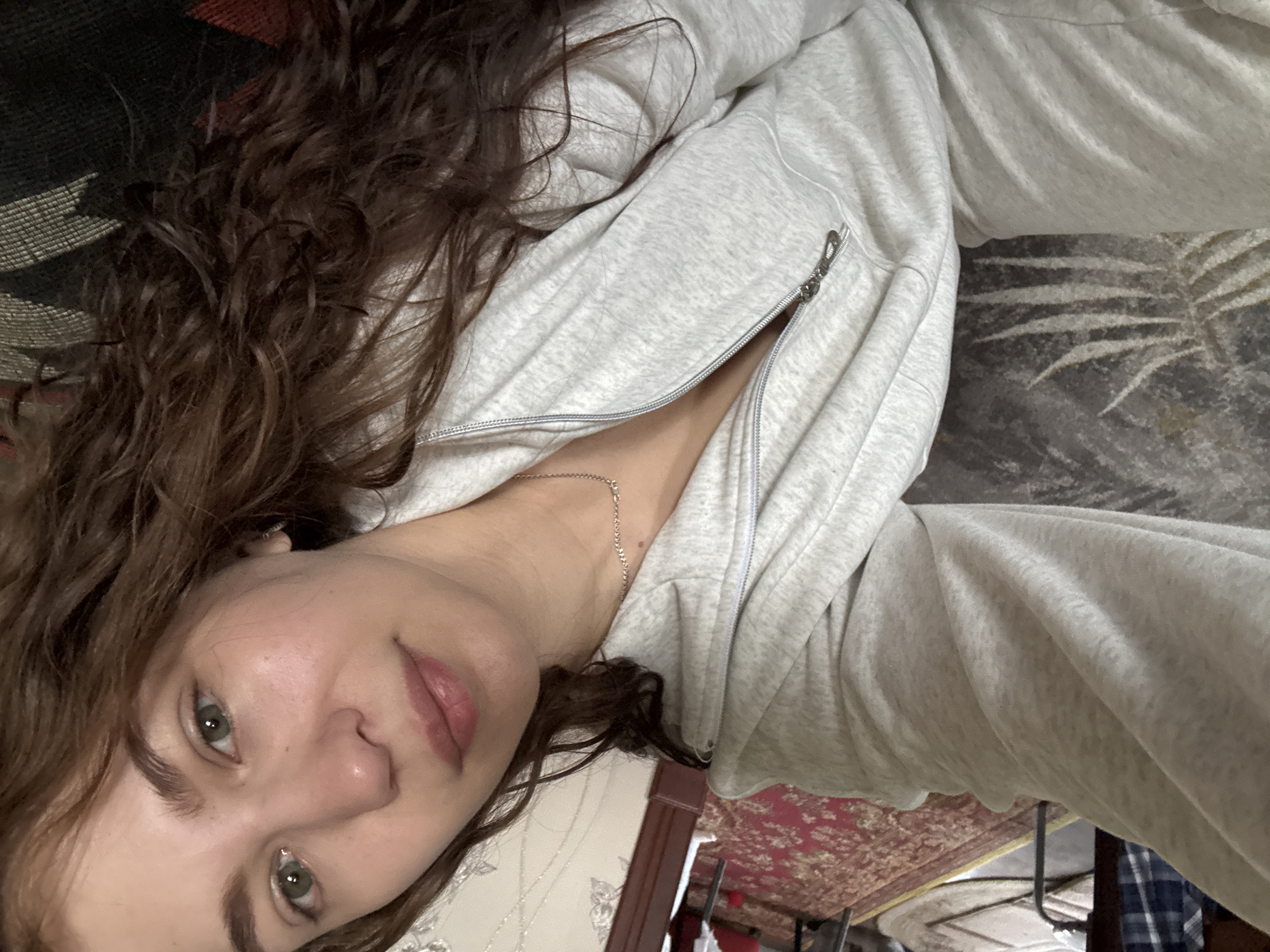 A woman with light skin, light green eyes, and wavy brown hair is lying down. She is wearing a light gray zippered hooded sweatshirt and a silver necklace. A piercing is visible on the left side of her nose. The background includes patterned rugs in shades of red, black, and gray. Furniture legs are also visible. It is not possible to confirm the specific location in Nizhnyaya Gar', Russia from the visual information provided.