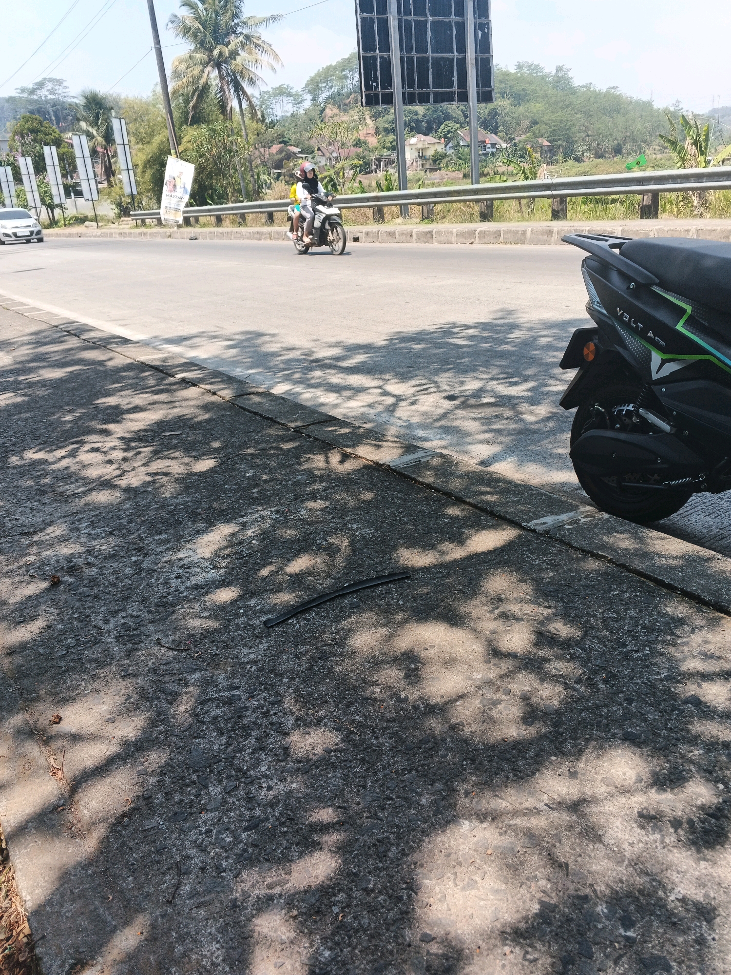 In Kalirejo, Indonesia, a street scene unfolds under a bright sky. A motorcycle carrying two individuals travels on an asphalt road, bordered by a metal guardrail. On the left, a white car is visible. The foreground is a concrete sidewalk, dappled with the shadows of trees. A dark, flexible object lies on the sidewalk. In the background, lush green hills dotted with houses rise towards the sky. A tall pole with posters stands on the left side of the road, and a large black structure, possibly a billboard or speaker, is present in the upper right corner.