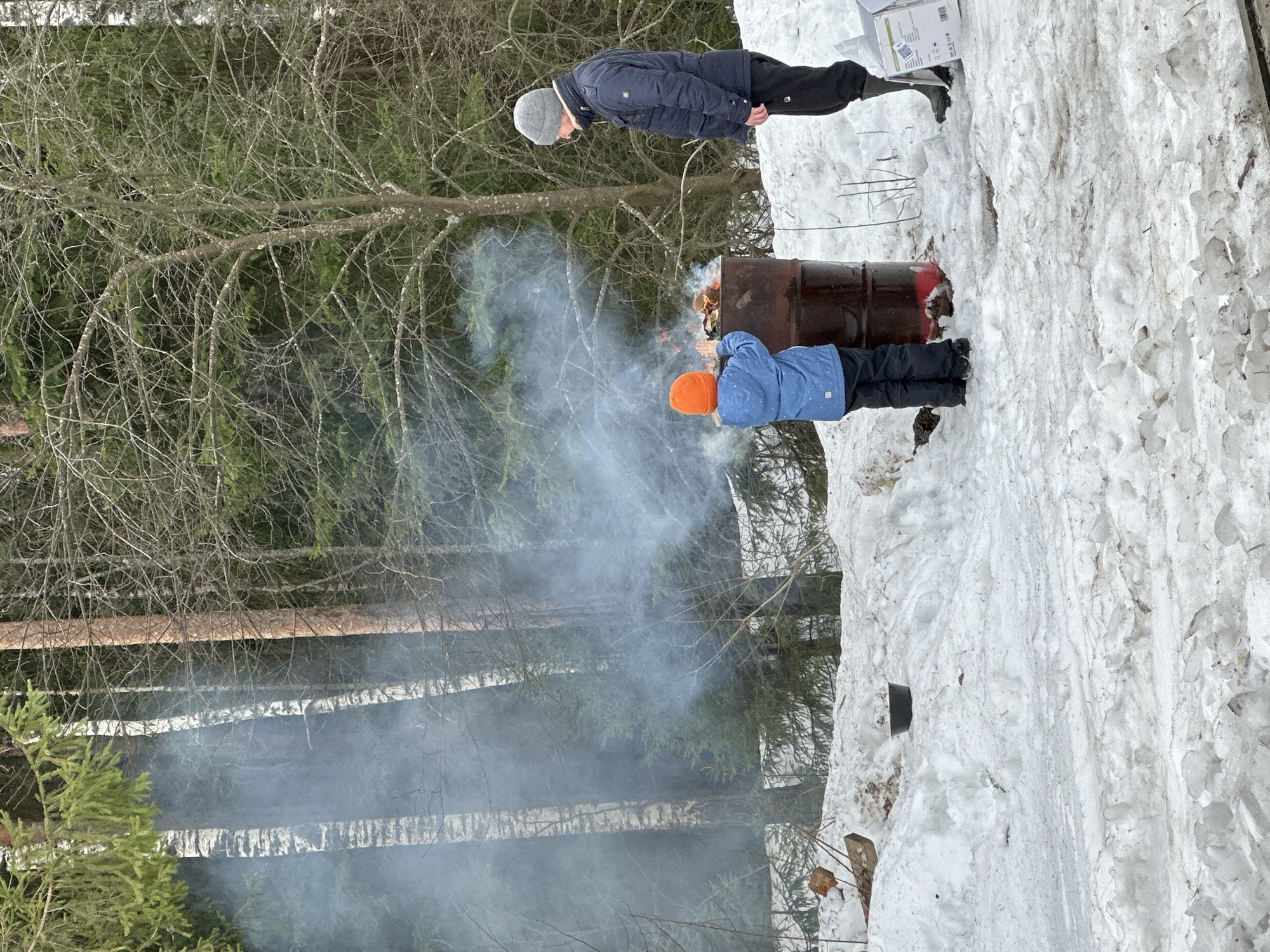 The image depicts a snowy, wooded area in Прикамский, Russia. Two individuals are standing near a metal barrel, which appears to contain a fire, producing a large plume of smoke. One person wears a blue jacket and orange hat, appearing to tend to the fire. The other person is wearing a black coat and gray hat. A box sits on the snow nearby. The ground is covered in snow, and trees are visible in the background.