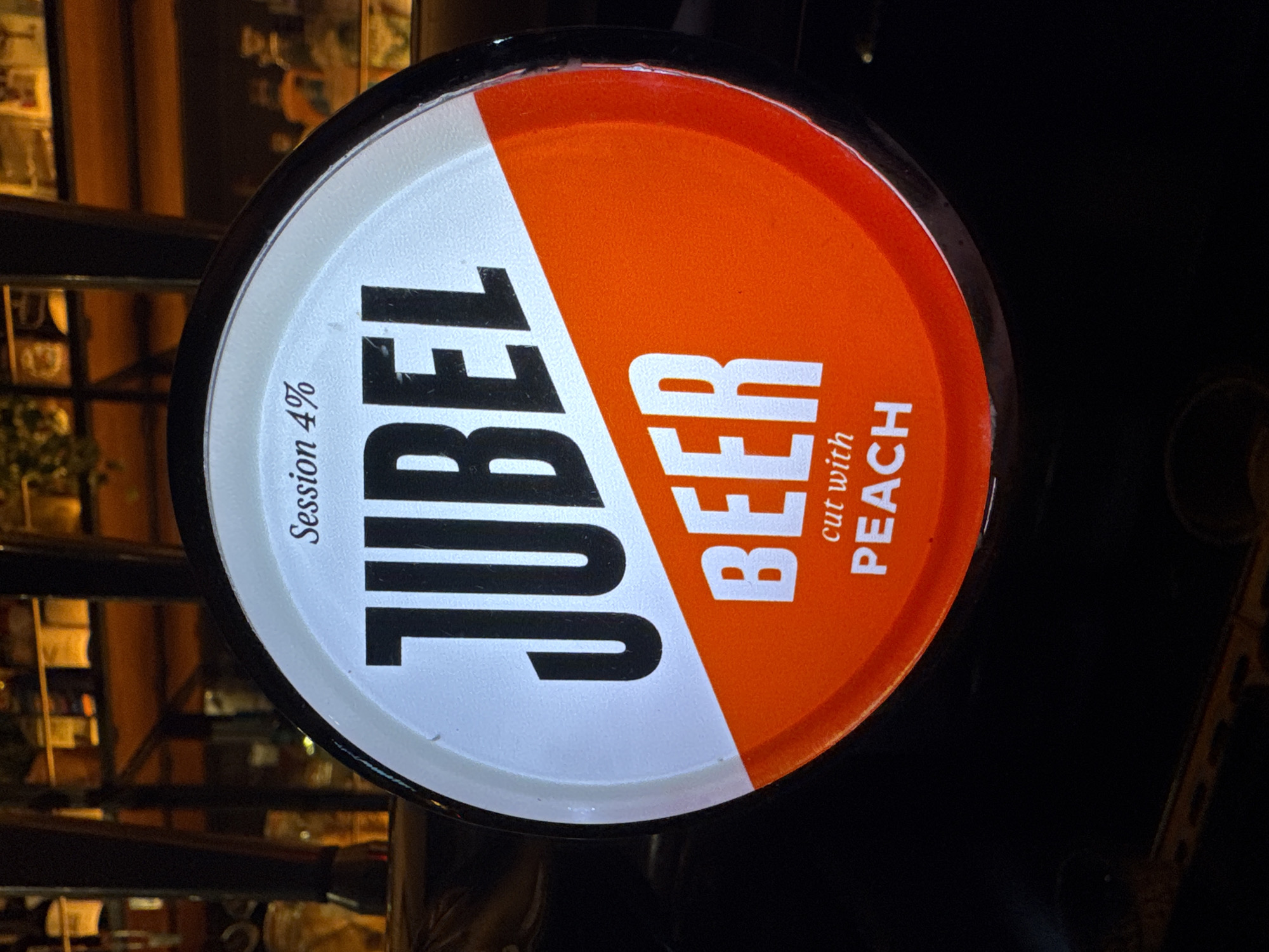 The image shows a Jubel beer tap handle in a dimly lit pub setting, likely in London, United Kingdom. The tap handle is circular, split diagonally into white and orange sections. "JUBEL" is vertically oriented in bold, black letters on the white section, with "Session 4%" above. On the orange section, "BEER" is vertically oriented in bold, white letters, with "cut with PEACH" below. The background features blurred glimpses of the pub interior, including shelves and other patrons. The light is low and warm, typical of pub settings.