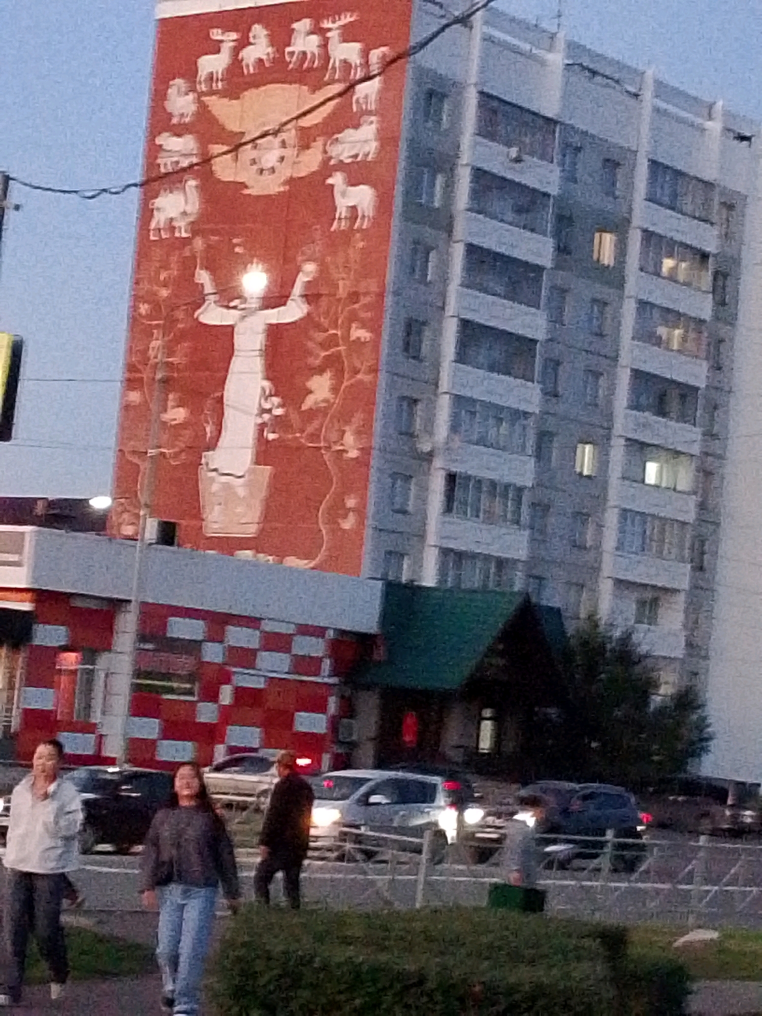The image shows a tall, multi-story building in Kyzyl, Russia, featuring a large mural on its side depicting animals and a central human-like figure holding an illuminated object. The building's facade is predominantly light gray, with numerous windows. Adjacent to this building, a lower structure with a dark green roof is visible. In the foreground, a street scene unfolds with several individuals walking and cars on the road. The lighting suggests it is either dusk or early morning, with some artificial lights from the building and street visible. The overall atmosphere is urban, with elements of public art and everyday street life.