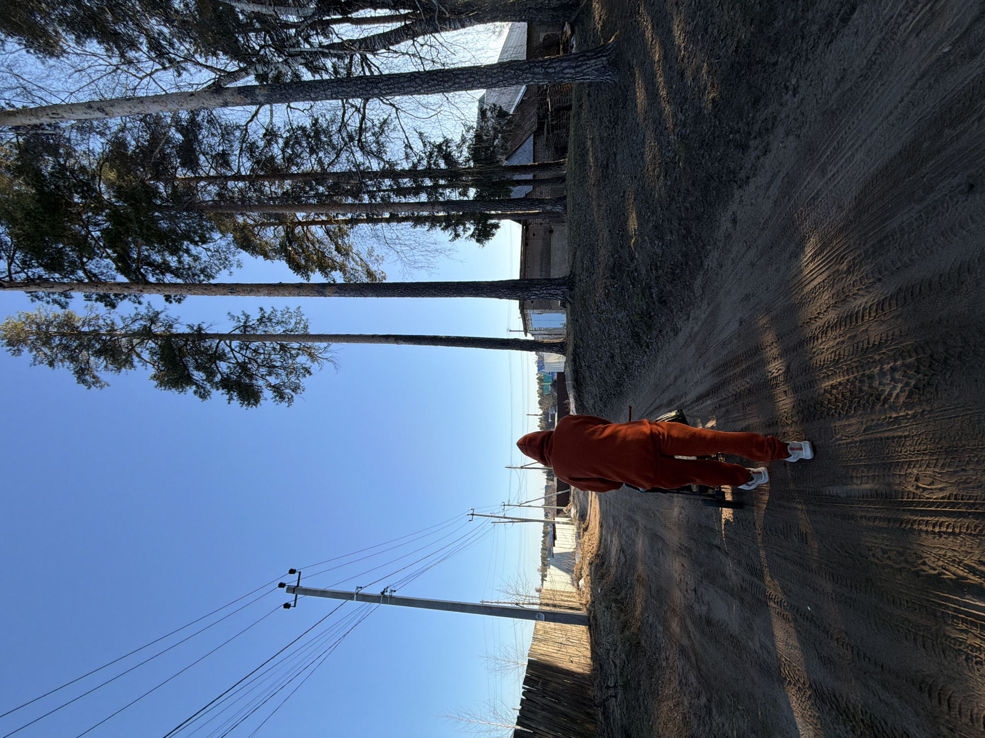 The image captures a rural or semi-rural scene along a dirt road in Chaikovsky, Russia, under a clear, bright blue sky. The perspective is from slightly behind a person, looking down the road into the distance.
The main subject is a person, seen from the back, wearing a vibrant orange hooded tracksuit and white sneakers. They are positioned on the right side of the dirt road, which is dry, dusty, and marked with numerous tire tracks. A small, dark object, possibly part of a bike or cart, is partially visible near their legs. Long shadows, likely cast by the trees and utility poles, stretch across the road, suggesting it is either early morning or late afternoon.
To the left of the road, a line of tall, slender coniferous trees, possibly pines, stands against the sky. Some bare deciduous trees are also visible among them. To the right, several wooden utility poles with multiple power lines extend along the roadside, accompanied by a rustic wooden fence. In the background, glimpses of distant buildings and structures indicate a village or settlement, with a faint horizon visible beyond.
The weather appears sunny and clear. No visible text is present in the image. The overall impression is one of calm and open space.