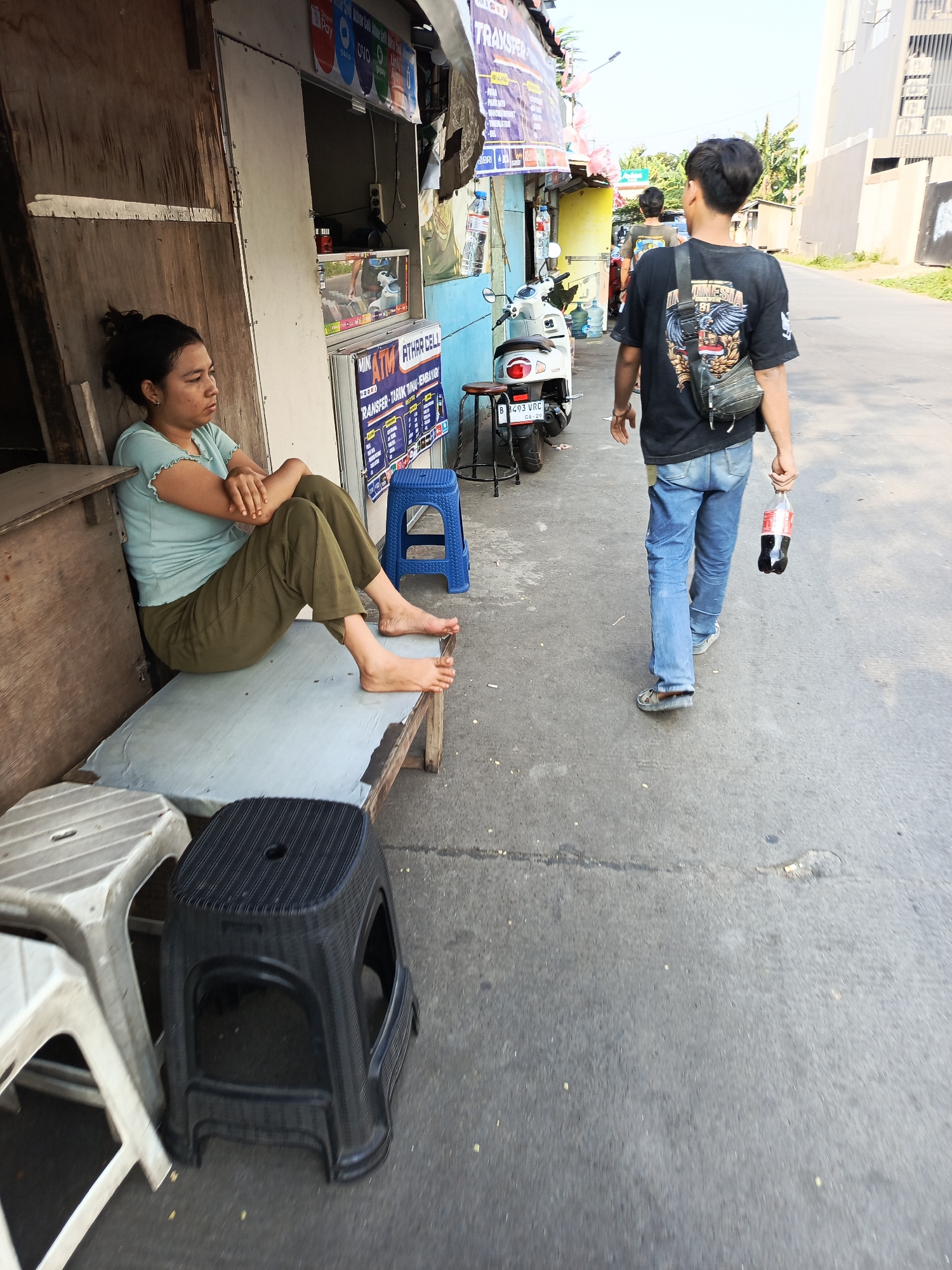 A young woman with her dark hair tied back sits on a wooden bench outside a shop in Tangerang, Indonesia. She wears a light blue short-sleeved shirt with ruffled edges and olive green pants. Her arms are crossed, and her bare feet are resting on the bench. Behind her, the shop has a light-colored sliding door and a sign with text in Indonesian. To her right, a young man in a black t-shirt with a graphic design on the back and blue jeans walks away from the camera, carrying a bottle of Coca-Cola in his right hand. He has a crossbody bag slung over his shoulder. In the foreground, there are several plastic stools, one white and two black. The scene is on a paved street with buildings and greenery visible in the background. The sky is clear and bright.