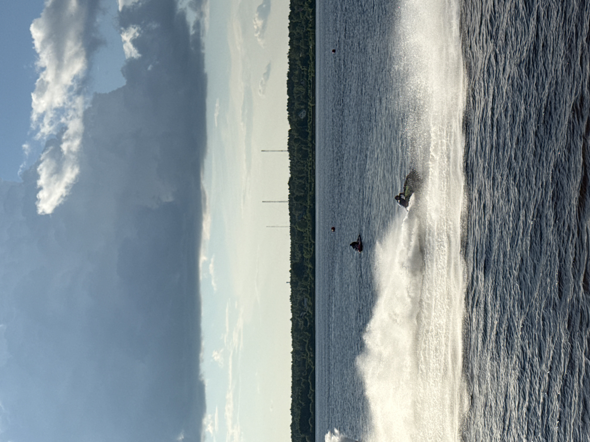 A vertical, wide-angle shot captures a dynamic scene on a body of water, likely a river or lake, with a treeline and structures on the horizon under a partly cloudy sky. In the foreground, dark blue, rippled water dominates the lower portion of the frame. The middle ground features a jet ski in motion, creating a large spray of white water as it moves from left to right. Further back, another jet ski is visible, also kicking up a smaller spray. A red buoy floats in the distance. The background shows a dense, dark green treeline along the bank of the water, with a few faint, tall structures, possibly communication towers or smokestacks, visible above the trees on the horizon. The sky is a mix of light blue with scattered white and dark gray clouds, suggesting potentially changing weather conditions. The overall impression is one of outdoor recreation on a waterway. The presence of tall, spindly structures in the background is consistent with industrial or infrastructural elements found in or near cities. Given the mention of the city of Perm, Russia, the scene likely depicts a location on the Kama River or one of its tributaries within or near the city's urban or suburban perimeter.