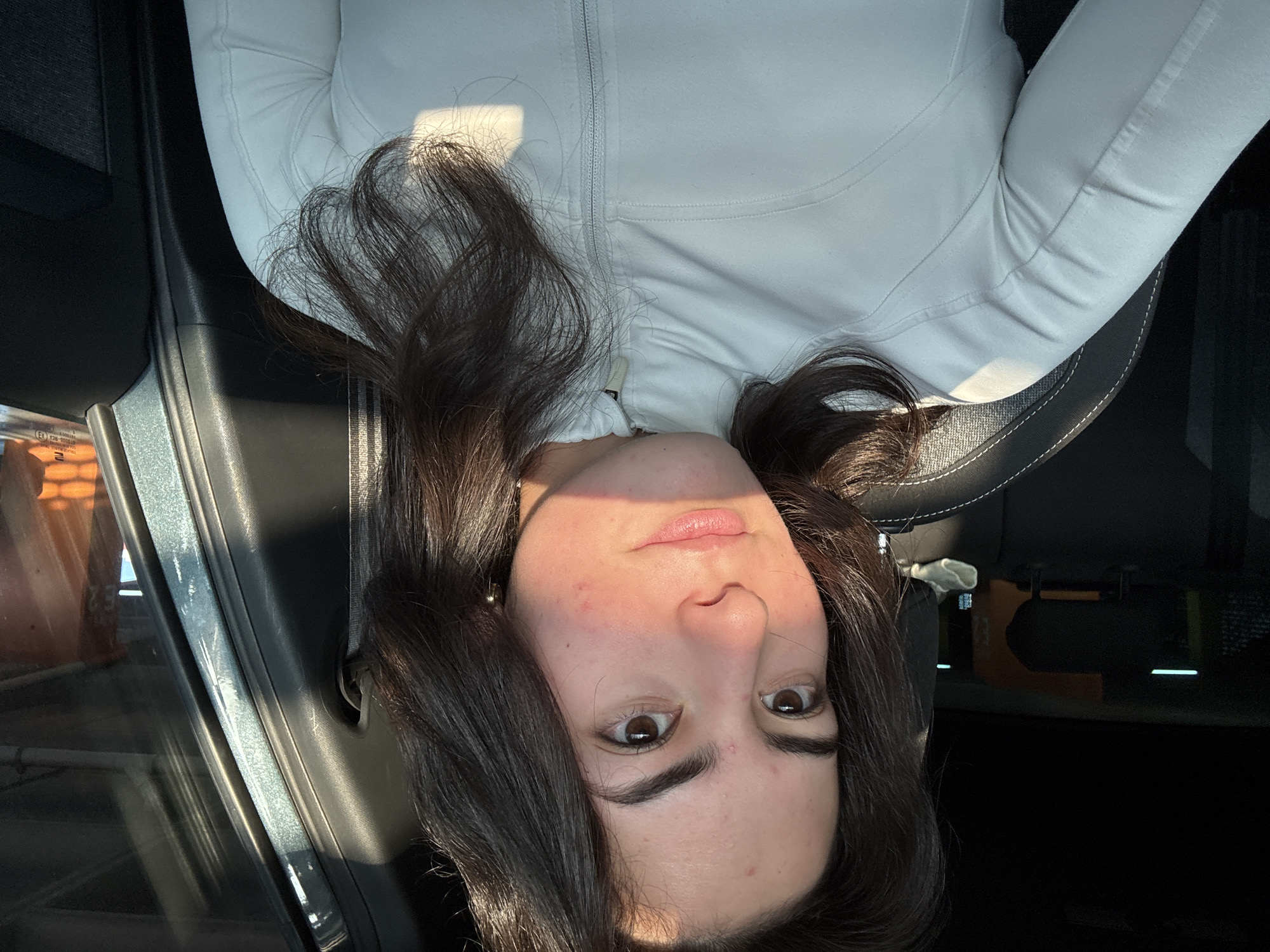 The primary image is an inverted, close-up portrait of a young woman with long, dark, wavy hair, taken inside a car. She is wearing a light-colored, possibly white or off-white, zippered athletic jacket. Her face is directed upwards, looking directly at the camera with a neutral, perhaps curious or pensive, expression. Her large brown eyes are open, and her lips are slightly parted. Strong, direct sunlight casts bright highlights across her forehead, nose, and lips, and illuminates strands of her hair. Some minor skin redness or blemishes are visible on her cheeks and forehead. A small, spherical earring is visible on her left ear.
The setting is the interior of a vehicle, evidenced by the car seat and headrest behind her. To the left, a car window and door frame are visible, through which bright daylight streams. Blurred elements outside the window suggest buildings or structures, placing the scene in an urban or semi-urban area, consistent with San Bernardino Tlaxcalancingo, Mexico. The intense, directional light indicates it is daytime, likely morning or late afternoon.
The woman appears to be reclining or leaning back in the car seat, with the camera positioned at a low angle. Her jacket features distinct seams and a central zipper. One cropped image reveals a faint label on the car's window or pillar that includes an "E" followed by numbers (e.g., "030"), likely indicating a regulatory marking. No other distinct activity is depicted beyond the woman looking at the camera.