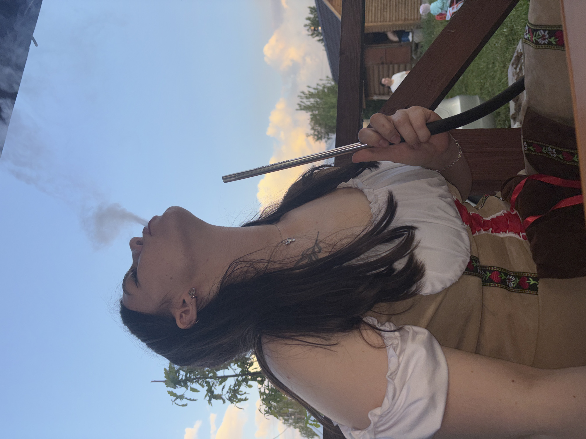 A woman is smoking a hookah outdoors in Пальники, Russia. She is wearing a traditional white and brown embroidered dress. Smoke emanates from her mouth as she holds the metal hookah pipe. The sky is visible in the background with some light clouds. Parts of a wooden structure are also visible. Other people can be seen in the background, possibly sitting at tables.
