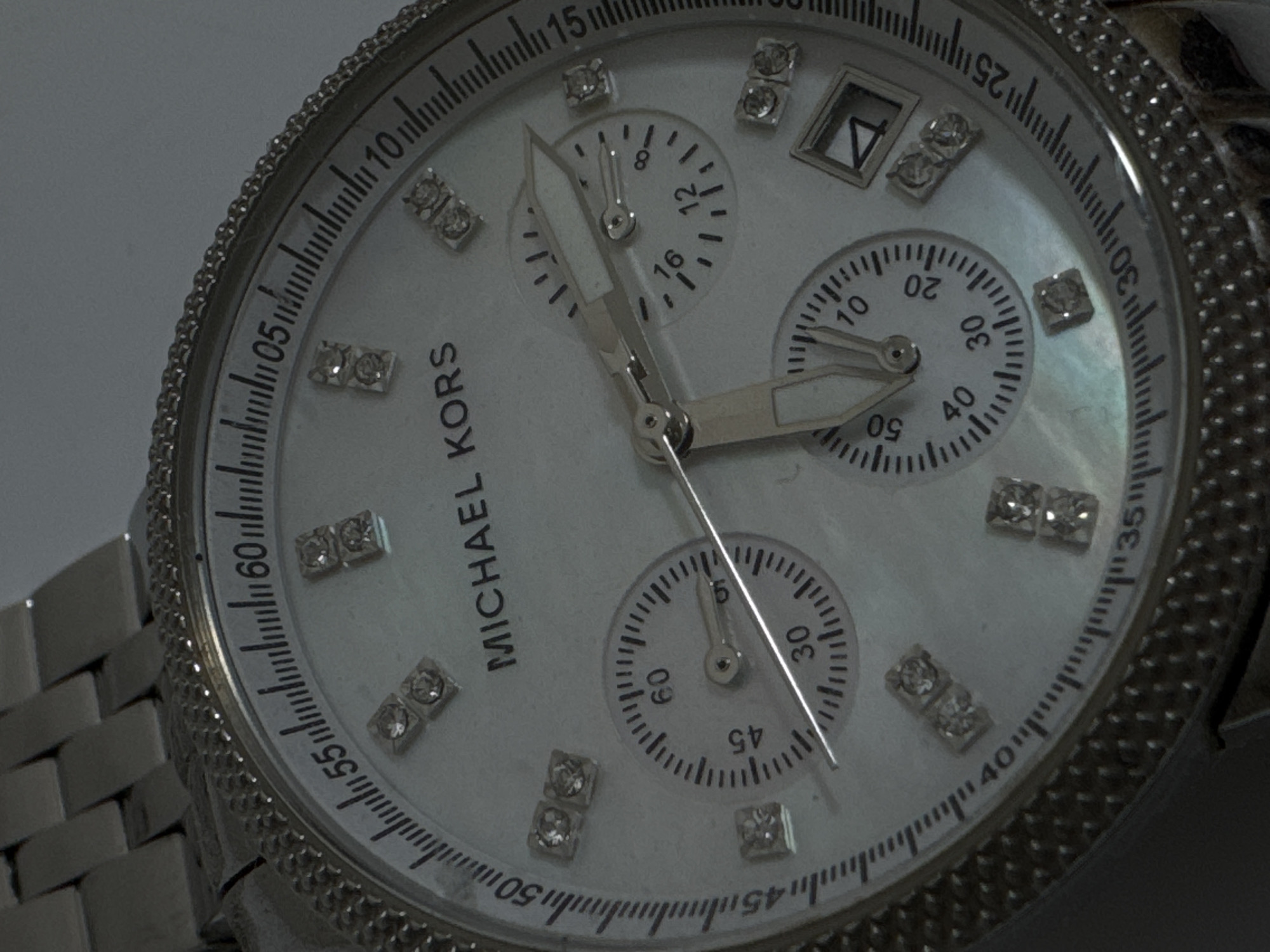 The image shows a close-up, angled shot of a silver Michael Kors watch. The watch has a white mother-of-pearl dial, with diamond hour markers. There are three sub-dials on the watch face. The outer bezel of the watch is encrusted with small, clear stones, likely diamonds, and the band is also silver. The lighting in the image is dim, causing some glare on the watch face and crystal. The watch is a chronograph model. There are no people or any indication of a city or location visible in the image.