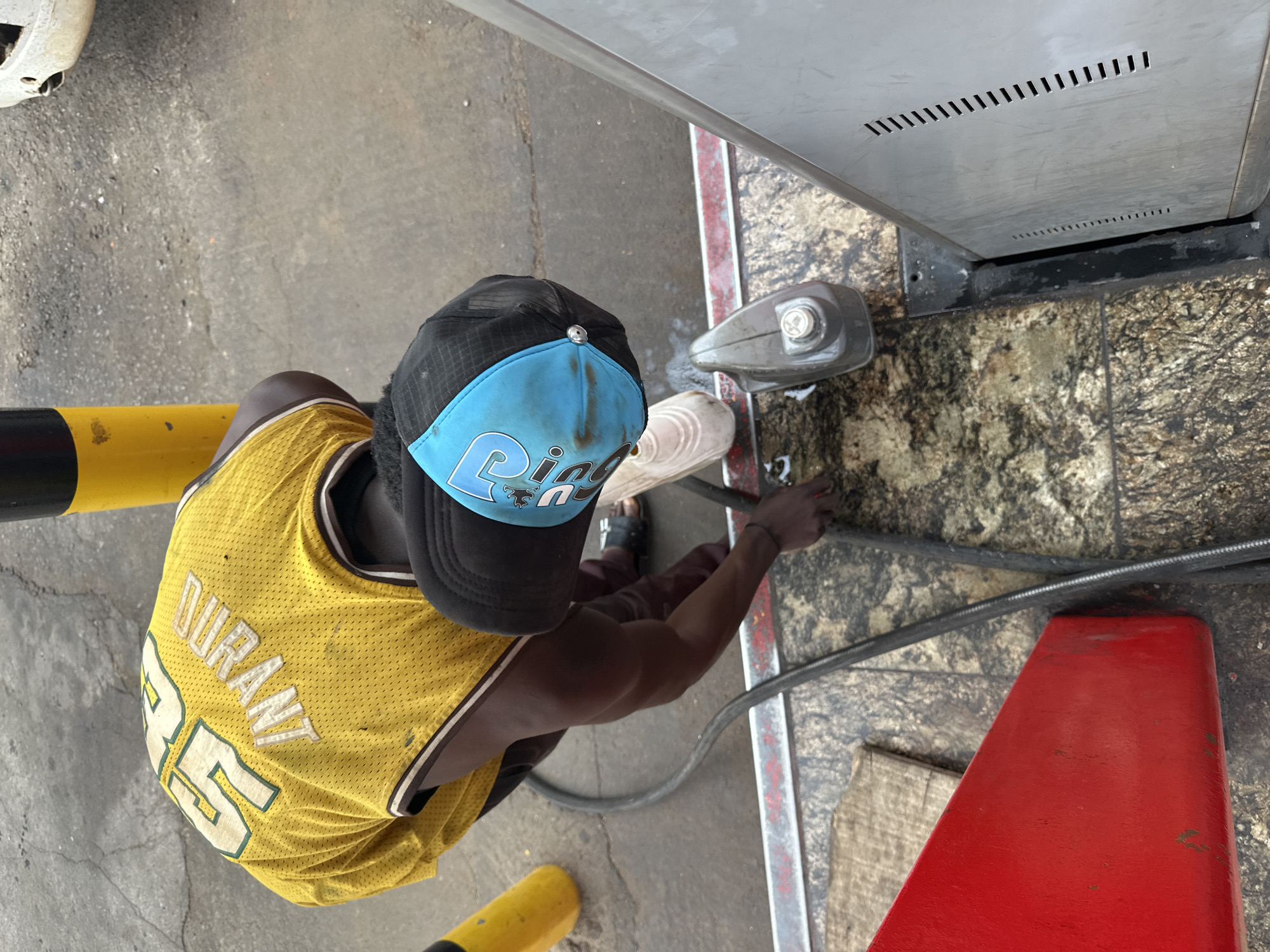 A high-angle shot shows a man in a yellow Durant #35 jersey, a black and light blue baseball cap, and sneakers standing at a metal fuel pump at a gas station. He is facing the pump and appears to be operating it. The ground is concrete with some staining. A yellow and black bollard is visible to the left of the man. A red safety cone is to the right of the man. The image was taken in the city of Jos, Nigeria.