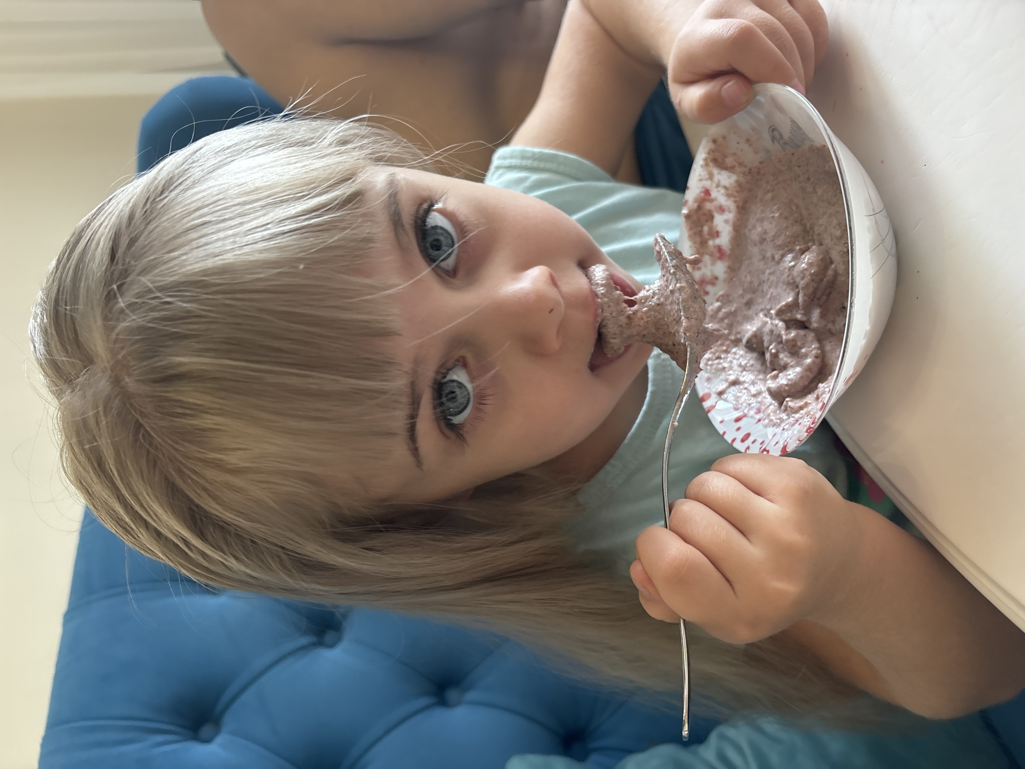 A young child with blond hair and blue eyes is shown eating from a bowl. The child is holding a silver fork with a dollop of brown substance on it, which she is putting into her mouth. The bowl appears to contain the same brown substance. The child is wearing a light blue shirt. Part of a blue, tufted cushion is visible in the background, and a white surface is next to the bowl. The location is Unknown, Unknown.