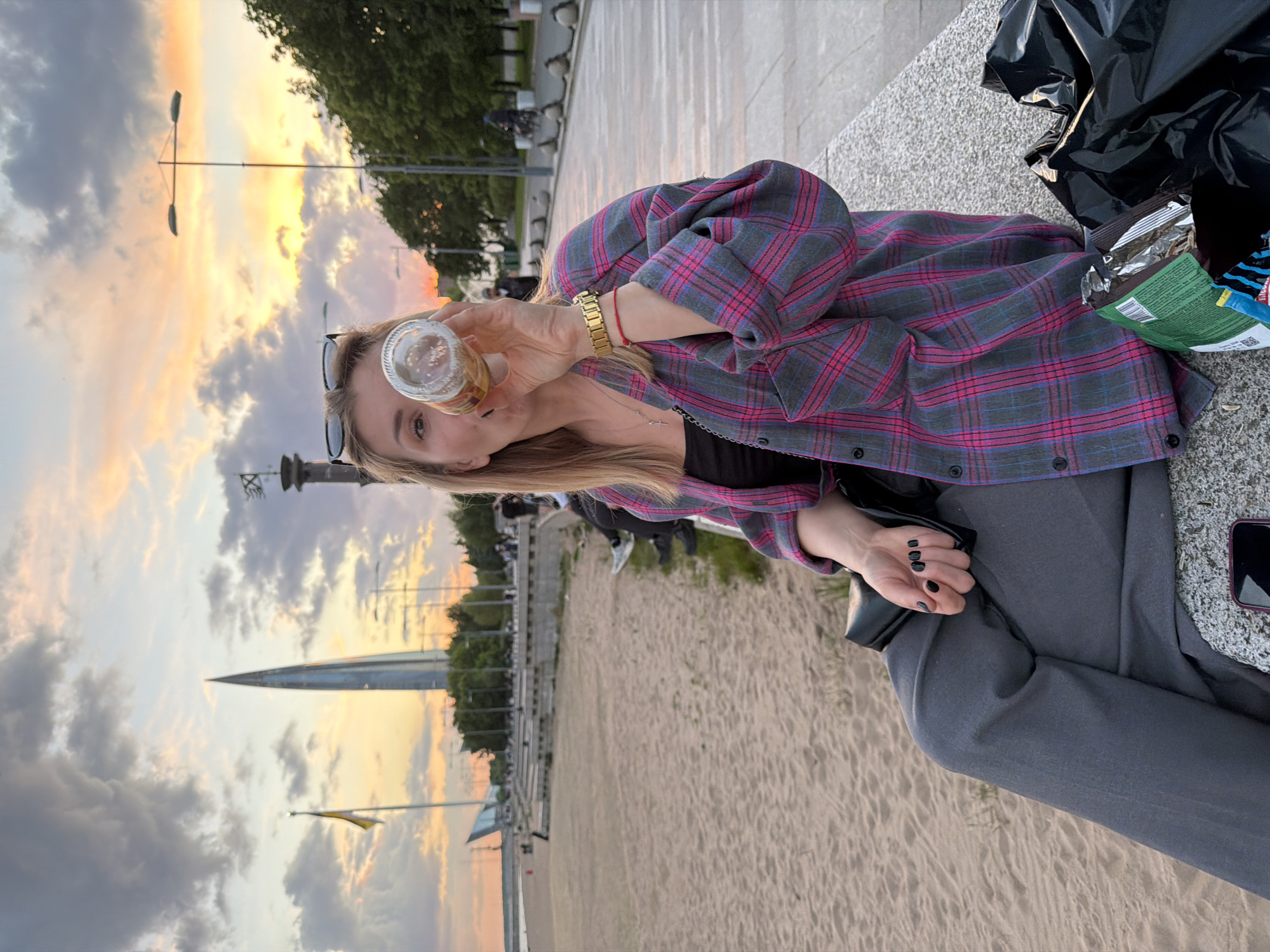 The image captures a young woman seated casually on a low, wide stone wall or bench during sunset or dusk, likely at a beach or riverside promenade in Saint Petersburg, Russia. She is looking directly at the viewer with a slight smile, holding a small, clear glass bottle or jar with a drink in her right hand, positioned near her face.
The woman has blonde hair, with sunglasses pushed up onto her head. She is dressed in a grey and pink plaid button-up shirt worn over a dark top, paired with dark grey trousers. Notable accessories include a gold-toned watch on her left wrist, a thin red string bracelet, and black nail polish on her fingers.
Beside her, on the stone surface, there is a black plastic trash bag and an open bag of snacks with some unreadable text visible. A smartphone is also partially visible near her leg.
The background showcases a beautiful sunset sky, filled with clouds displaying warm hues of orange, pink, and purple against a fading blue. Behind the woman, a sandy area suggests a beach or riverbank, extending towards a distant cityscape. Further back, streetlights, trees, and a prominent, tall, spire-like architectural structure are visible against the horizon. The setting evokes a serene and relaxed evening atmosphere.