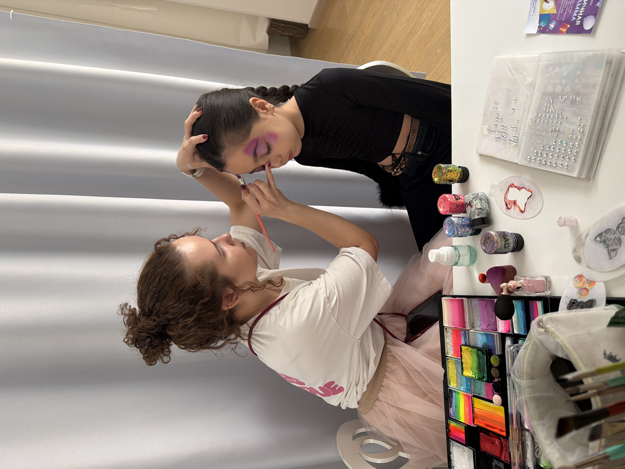 This indoor photograph captures two young females engaged in face painting or artistic makeup application. One person, with curly brown hair tied in a bun, is applying intricate makeup to the other. She wears a light-colored t-shirt with partially visible pink text that reads "LOVE" and a light pink tulle skirt. The subject receiving the makeup has dark hair styled in a braid and is wearing a black long-sleeved top and dark pants with a decorative belt. Her eyes are closed, revealing vibrant purple and pink eyeshadow or face paint already applied. She also has red nail polish.
The activity appears to be taking place in front of a light grey pleated curtain or backdrop. To the right, a white table holds a comprehensive collection of face painting and makeup supplies. These include a large palette filled with various brightly colored paints, some arranged in rainbow gradients. Several pots of glitter in gold, red, silver, blue, and purple are visible, with one black-capped pot clearly labeled "MAC". Sheets of stick-on rhinestones or gems, various brushes, and small bottles of liquids are also scattered on the table. Two stencils, one resembling a tooth and another a butterfly, are present. A small package on the table bears the Cyrillic text "ЯРКАЯ ГАММА" (Bright Palette) and "МАММА" (Mama). A light wooden floor is visible in the upper left corner of the image. The scene suggests a focused and calm atmosphere, likely an artistic session or preparation for an event, located in Saint Petersburg, Russia.