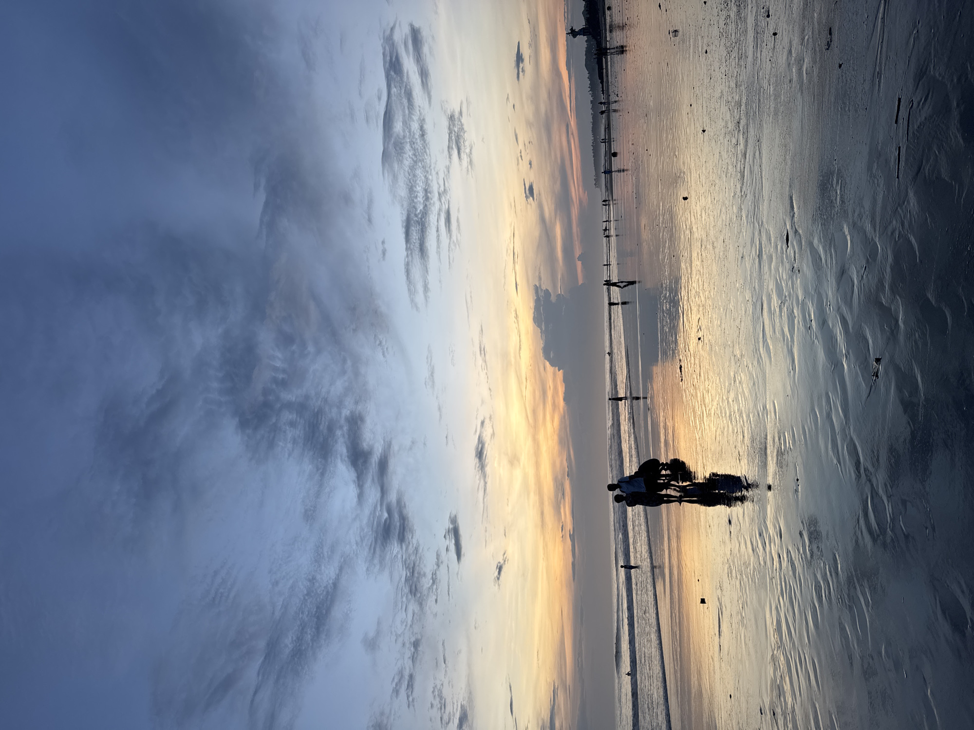 The image depicts a beach scene at sunset in North Kuta, Indonesia. The sky exhibits a gradient of colors, ranging from dark blue with striated clouds to a band of orange and yellow near the horizon. The foreground consists of wet sand reflecting the colors of the sky. Several silhouetted figures are visible on the beach, including a group of people gathered near the center of the frame. Individual figures are spaced along the water's edge, and distant structures are suggested on the horizon.