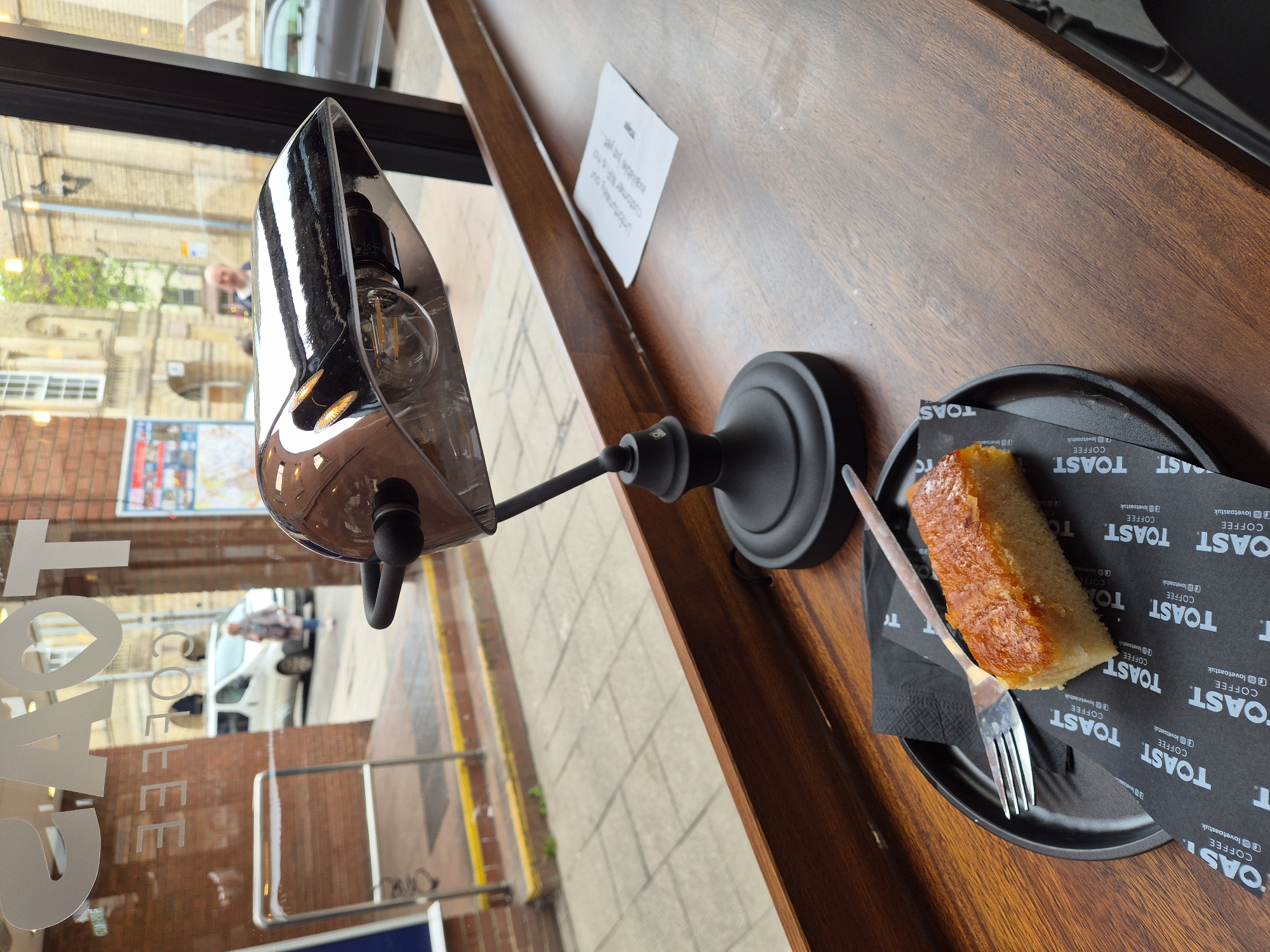 The image shows an indoor café scene in West Suffolk, United Kingdom, likely in Bury St Edmunds based on the view from the window. The camera angle is from a seated position, looking across a wooden counter. On the counter, there's a black plate with a slice of cake resting on black paper napkins printed with the words "TOAST COFFEE" and social media handles. A fork is next to the cake. Beside the plate, there is a black lamp with a transparent, rounded shade. There's also a white rectangular sign. The café window displays the words "TOAST COFFEE" in large, white letters. Reflected in the window and visible through it is a street scene, showing a building façade, a map, pedestrians, and vehicles. The overall impression is one of a modern café setting.