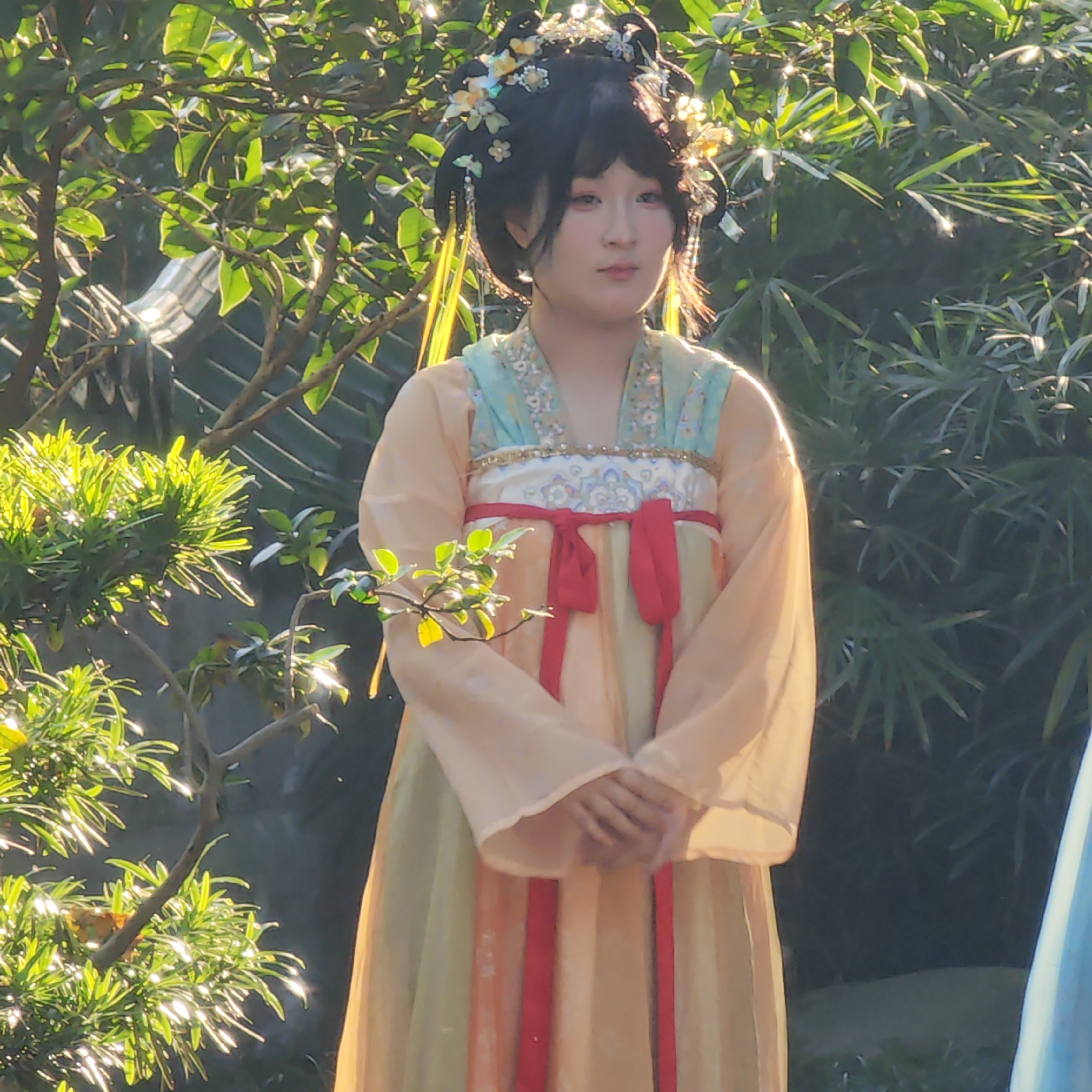 A young woman stands in a garden in the Yuzhong District of China, wearing a traditional Chinese Hanfu dress with peach-colored sleeves, a mint green collar, and a red sash. Her hair is styled in an elaborate updo adorned with flowers and ornamental hairpins. She is surrounded by lush green foliage, and sunlight dapples the scene. The architectural features visible in the background, such as a tiled roof, suggest a traditional Chinese garden setting. The woman has her hands clasped in front of her.