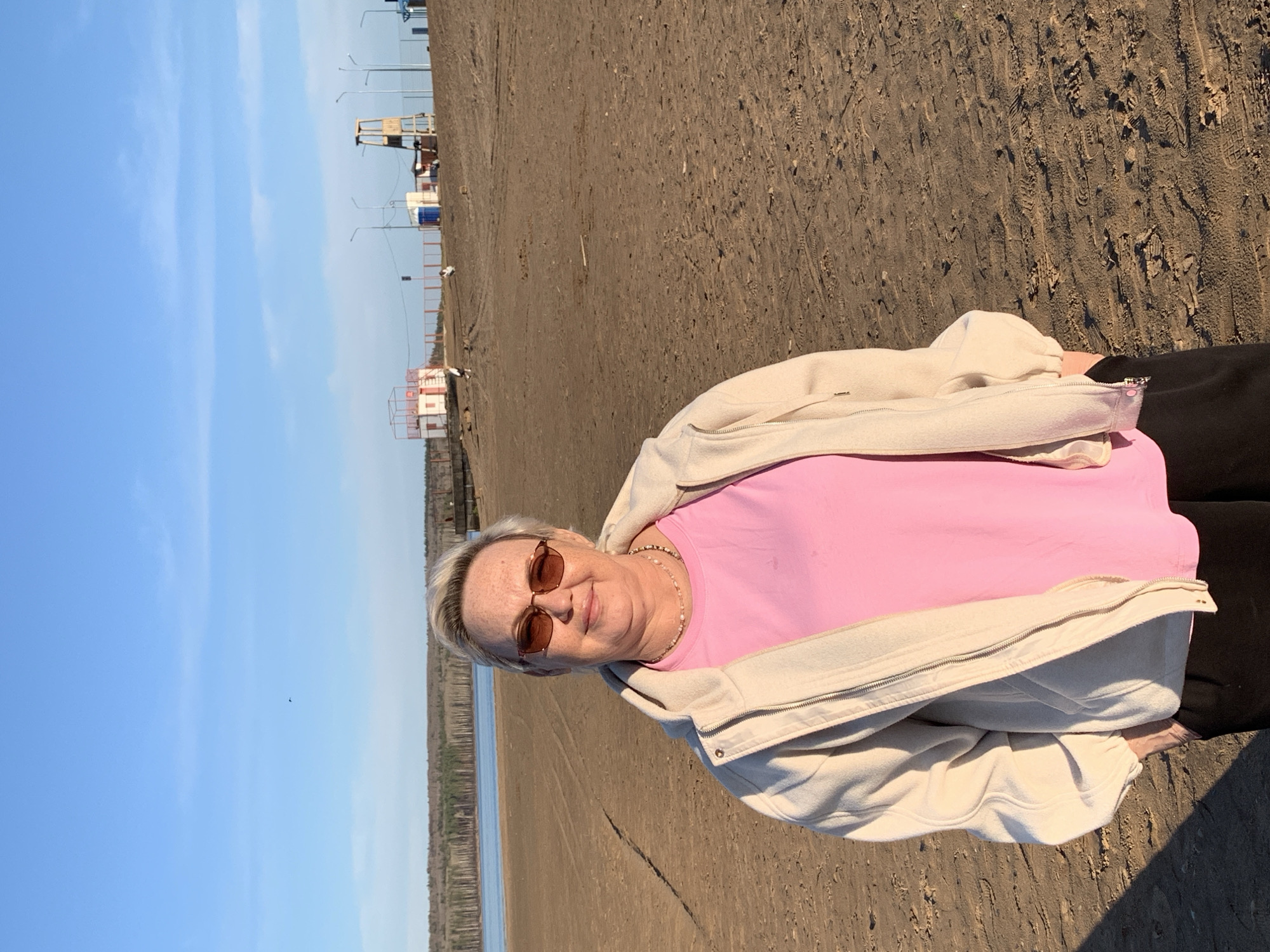 A woman stands on a beach, the city of Chaikovsky, Russia, is in the background. The woman has light-colored hair and sunglasses, wears a pink shirt, a light-colored jacket, and dark pants. The beach is brown and sandy, with many footprints. The sky is blue with a few clouds. In the background, a body of water is bordered by trees. Further back, structures and utility poles can be seen, indicative of the city.