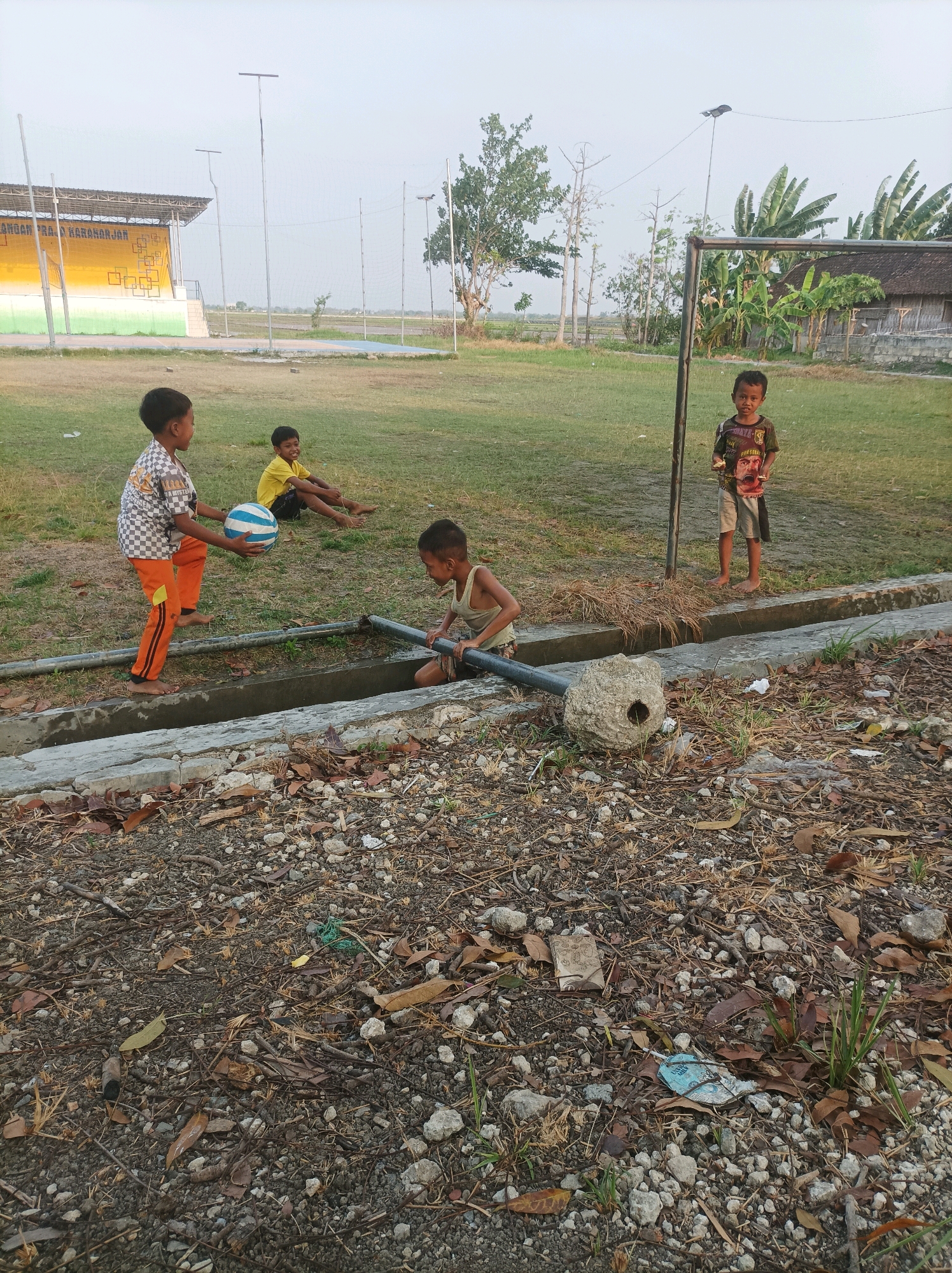 Four young boys are in a grassy outdoor area in SARANGAN, Indonesia. In the foreground, two boys are near a concrete drainage ditch with pipes. One boy, wearing an orange and black checkered shirt and orange pants, holds a blue and white striped ball. The other boy, wearing a light-colored tank top, is kneeling and holding a pipe that enters the ditch. In the mid-ground, a boy in a yellow shirt is sitting on the grass. Further back, near a soccer goal, another boy in a dark t-shirt with a graphic print and light shorts stands looking at the camera. Behind the children, there is a grassy field, a stadium structure with yellow and green panels, trees, and a traditional Indonesian house with a thatched roof. The ground in the immediate foreground is covered with soil, small stones, dry leaves, and twigs. The sky is overcast.