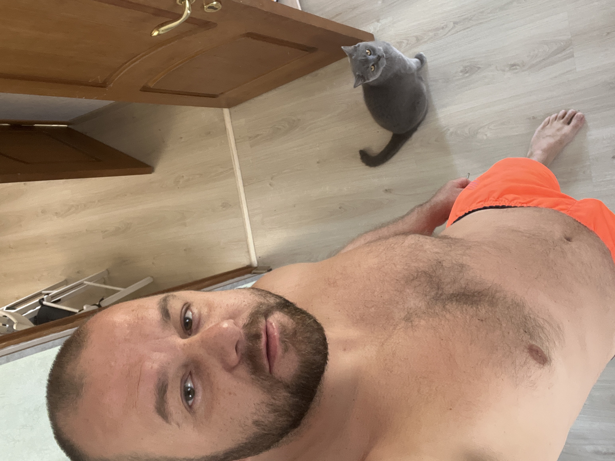 This image depicts an indoor scene, described as being located in Sochi, Russia. The primary subject is an adult male, likely Caucasian, captured in a high-angle selfie perspective, looking directly into the camera. He possesses short, dark, receding hair and a full, dark beard and mustache. His expression is neutral. The man is shirtless, revealing a moderately hairy chest and abdomen, and is wearing bright orange swim trunks or shorts with a visible black waistband. His right bare foot is visible on the light-colored floor.
To the man's right and slightly behind him on the floor is a grey (blue) British Shorthair cat, sitting upright with bright amber eyes, also looking towards the camera. Its tail is visible on the floor.
The environment features light-colored laminate or wooden flooring throughout. Two brown wooden doors are visible in the background: one fully closed on the right side of the frame with a gold handle, and another partially ajar in the center-left, leading to a darker adjacent room. Through the partially open door, a white, multi-tiered drying rack or shelving unit is discernible. The walls appear light-colored. The scene is evenly lit.