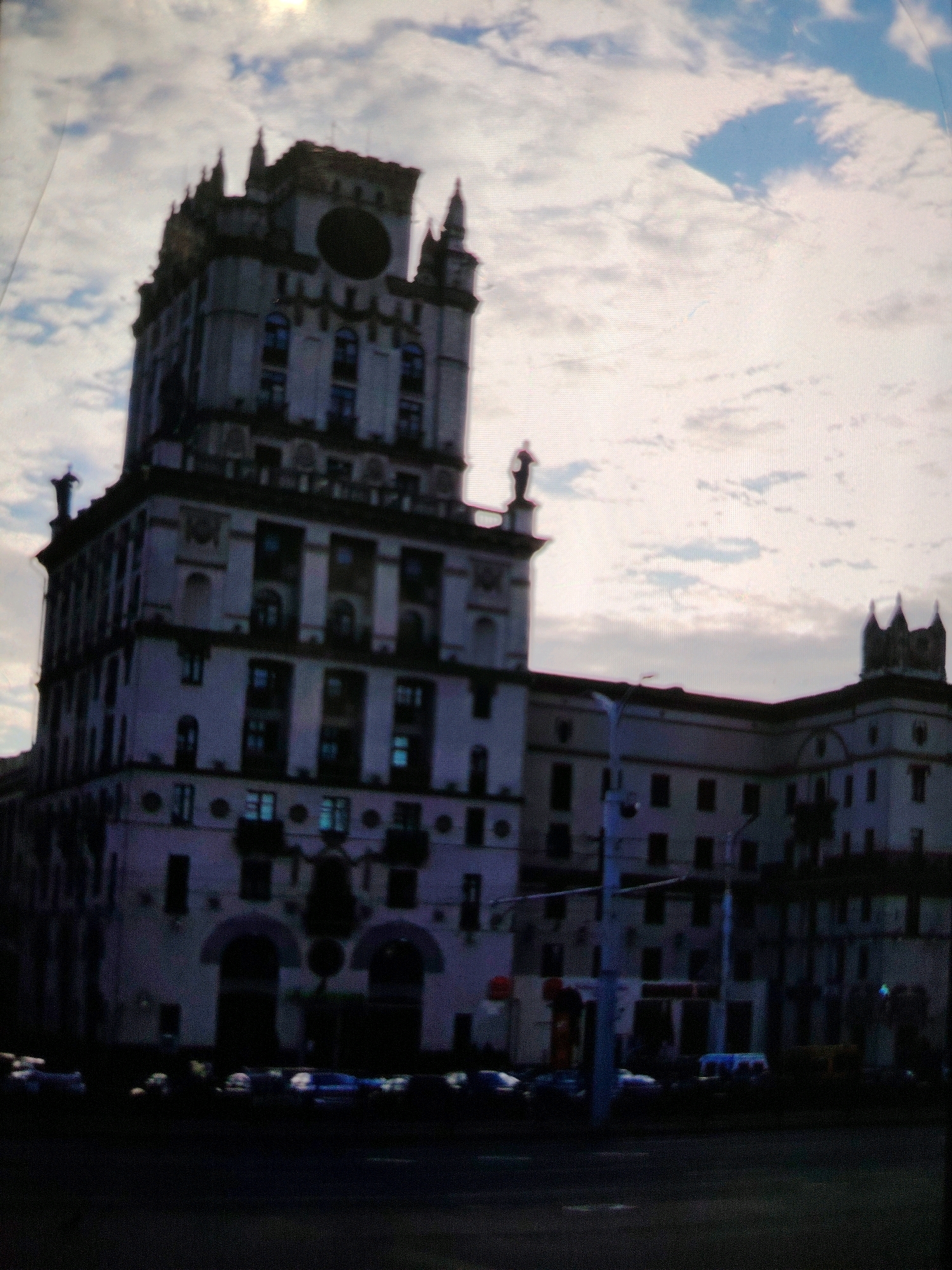 The image depicts the iconic Stalinist Empire style building, commonly known as the "Seven Winds" or "Gate of Mazyr," situated in Mazyr, Belarus. The building, a prominent landmark, is characterized by its imposing architecture, featuring multiple stories, decorative elements, and a central tower topped with spires. It stands against a backdrop of a cloudy sky, with a hint of blue peeking through. In the foreground, a street is visible with several vehicles parked or driving, suggesting urban activity. The lighting indicates it is either dusk or dawn, with the building illuminated by artificial lights and natural light from the sky. The overall scene captures the grandeur of the architecture and the everyday life of the city.